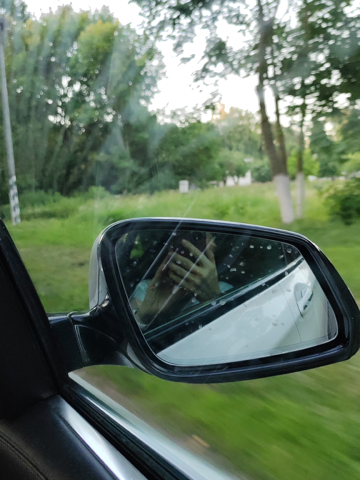 The image shows a reflection of a person's hands holding a smartphone in a car's side mirror. The mirror is slightly dirty with water droplets. The car is in motion, indicated by the blurred green landscape and trees visible through the car window and in the distance. The scene suggests the person is a passenger or driver, capturing a moment while traveling through a rural area. The location is identified as the Lyshchytsy Village Council, Belarus.