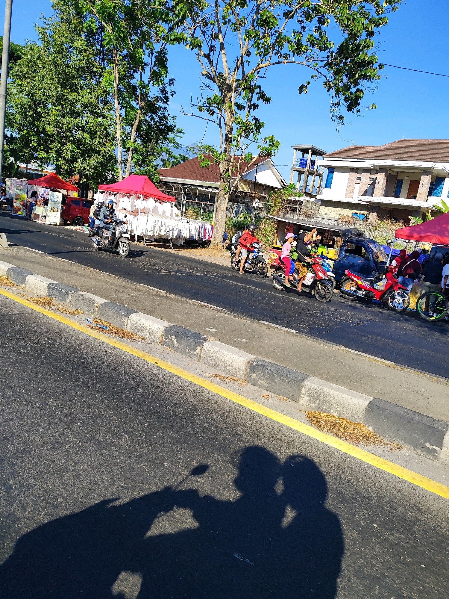 The image depicts a street scene in Salatiga, Indonesia, on a sunny day. In the foreground, the asphalt road is visible with a yellow line marking the edge of the lane and a concrete curb. The shadows of two people, likely on a motorbike, are cast on the road.
In the midground, several people are seen on and around motorbikes. Two individuals are riding a scooter, with the rider wearing a helmet and a blue jacket, and a passenger behind them. Further down the road, other motorbikes are visible, some with passengers. Several temporary stalls with red canopies are set up along the roadside, displaying various items, possibly clothing and food. A red car is parked near one of the stalls.
In the background, there are trees with green foliage and a clear blue sky. Several buildings with tiled roofs are also visible, suggesting a residential or commercial area. The overall scene suggests a bustling street with local commerce and daily life unfolding.