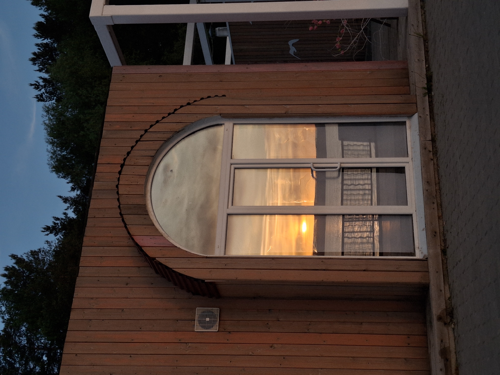The image depicts a section of a building exterior, likely in Chaikovsky, Russia, given the context provided. The building features wooden siding with a distinctive curved element, potentially part of a window frame or decorative architectural detail. A rectangular window with multiple panes is visible, reflecting a sunset or sunrise, indicated by the warm, golden light. The reflection suggests the presence of curtains or blinds inside the building. A small, square ventilation grate is present on the wooden siding below the window. To the left, tree foliage is discernible against a blue sky. The adjacent building is constructed of a gray stone brick. A porch structure is visible near the top of the image. There are no people depicted.