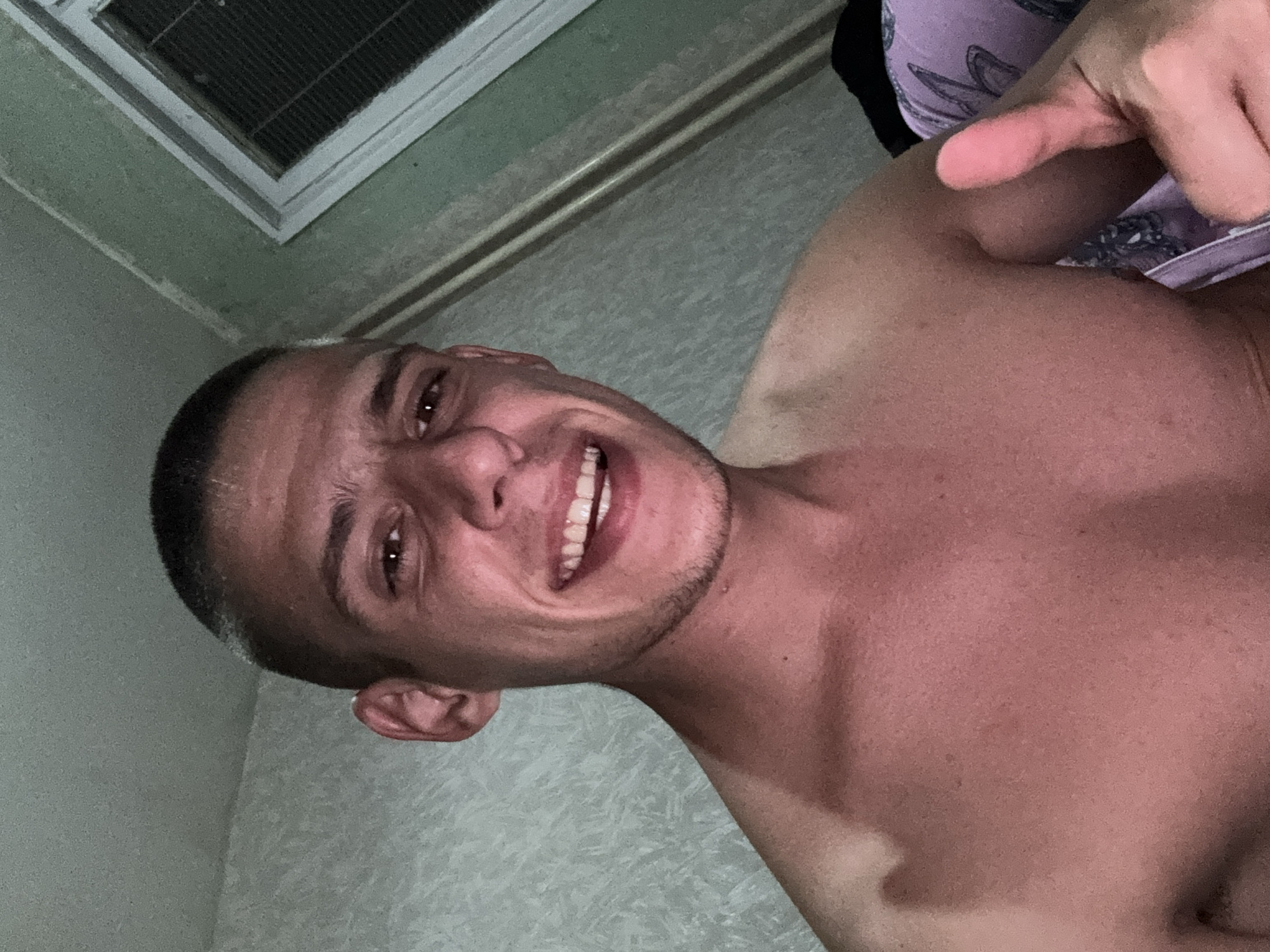 A man with short dark hair and a light complexion is smiling and giving a thumbs up gesture. He appears to be shirtless, with a patterned garment visible near his shoulder. The background is a pale wall with a vent near the ceiling. It is impossible to determine the specific location in Sofia, Bulgaria from this image alone.