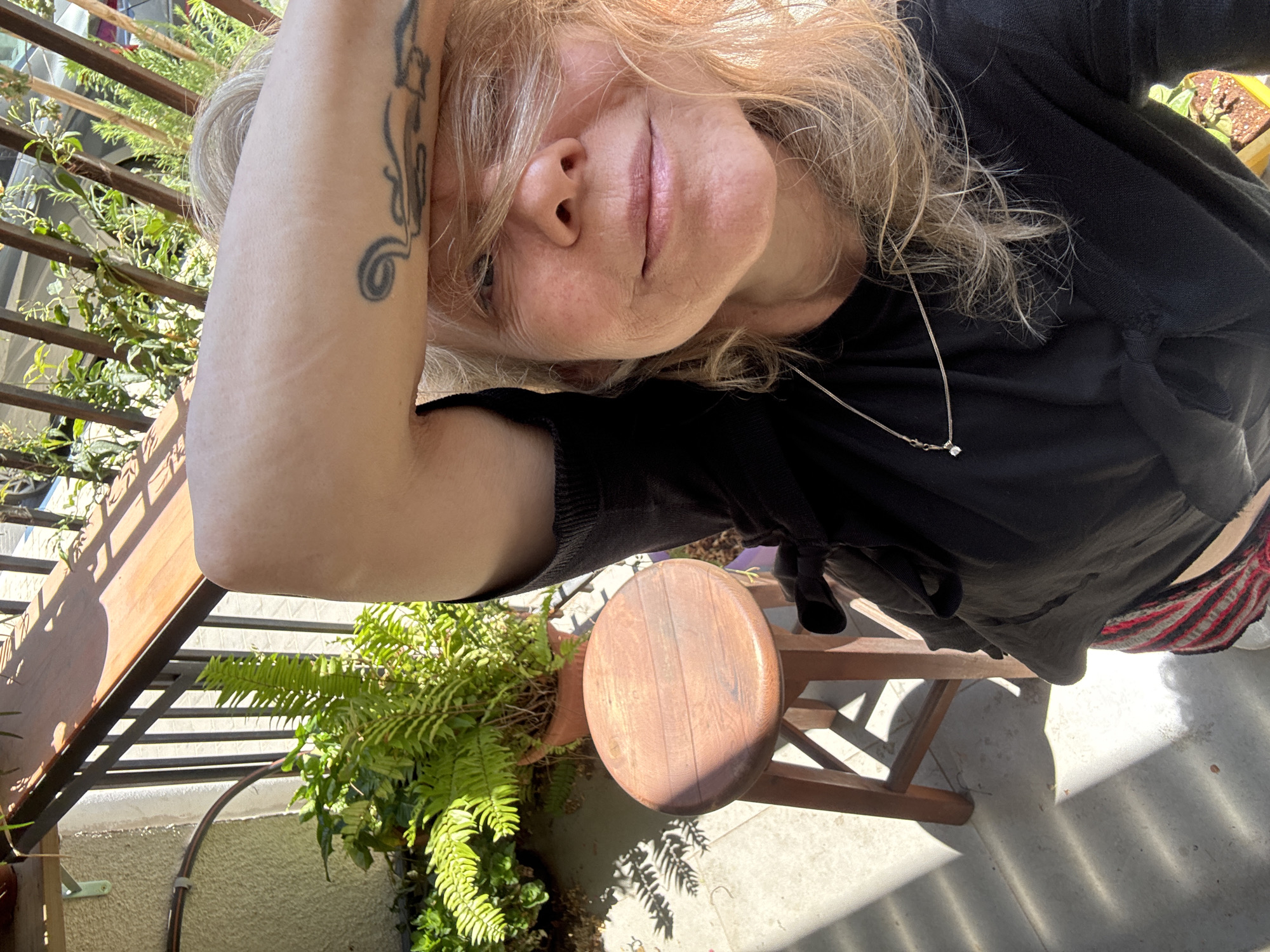 A woman with blonde hair and a tattoo on her arm is outdoors, possibly on a balcony or patio. She is wearing a black shirt and a necklace. There is a fern in a pot, a small wooden stool, and a wooden bench in the scene. The location is inferred to be Tel-Aviv, Israel, based on the background, foliage, and architecture which is not clearly visible.