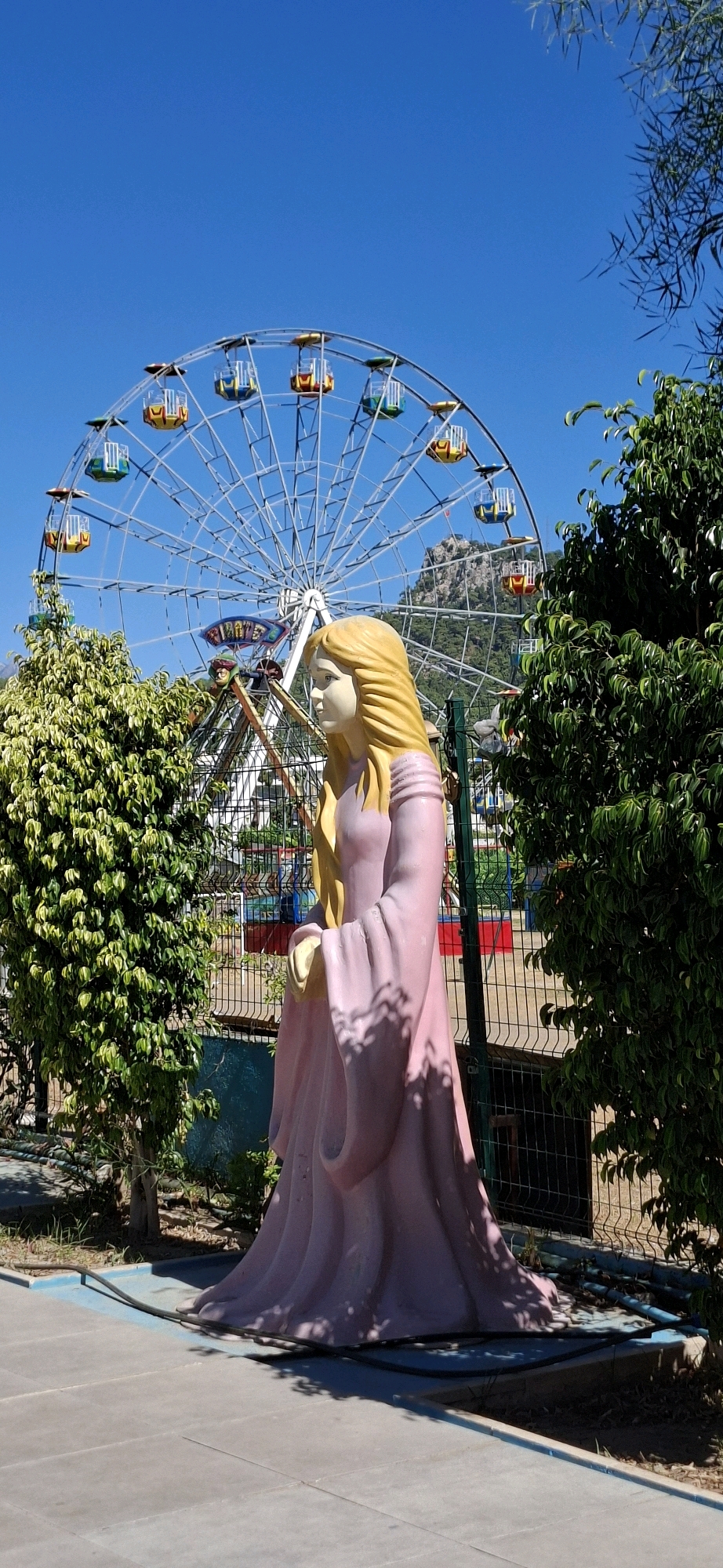 In the city of Kemer, Turkey, an amusement park features a ferris wheel, a statue of a blonde-haired woman in a pink dress, and lush green trees under a clear blue sky. The statue is positioned in the foreground, facing left, with the ferris wheel as a backdrop. The ferris wheel, a prominent attraction, has multi-colored gondolas and is set against a mountainous landscape. The surrounding area includes a metal fence, paved ground, and dense foliage, suggesting a warm, sunny day.