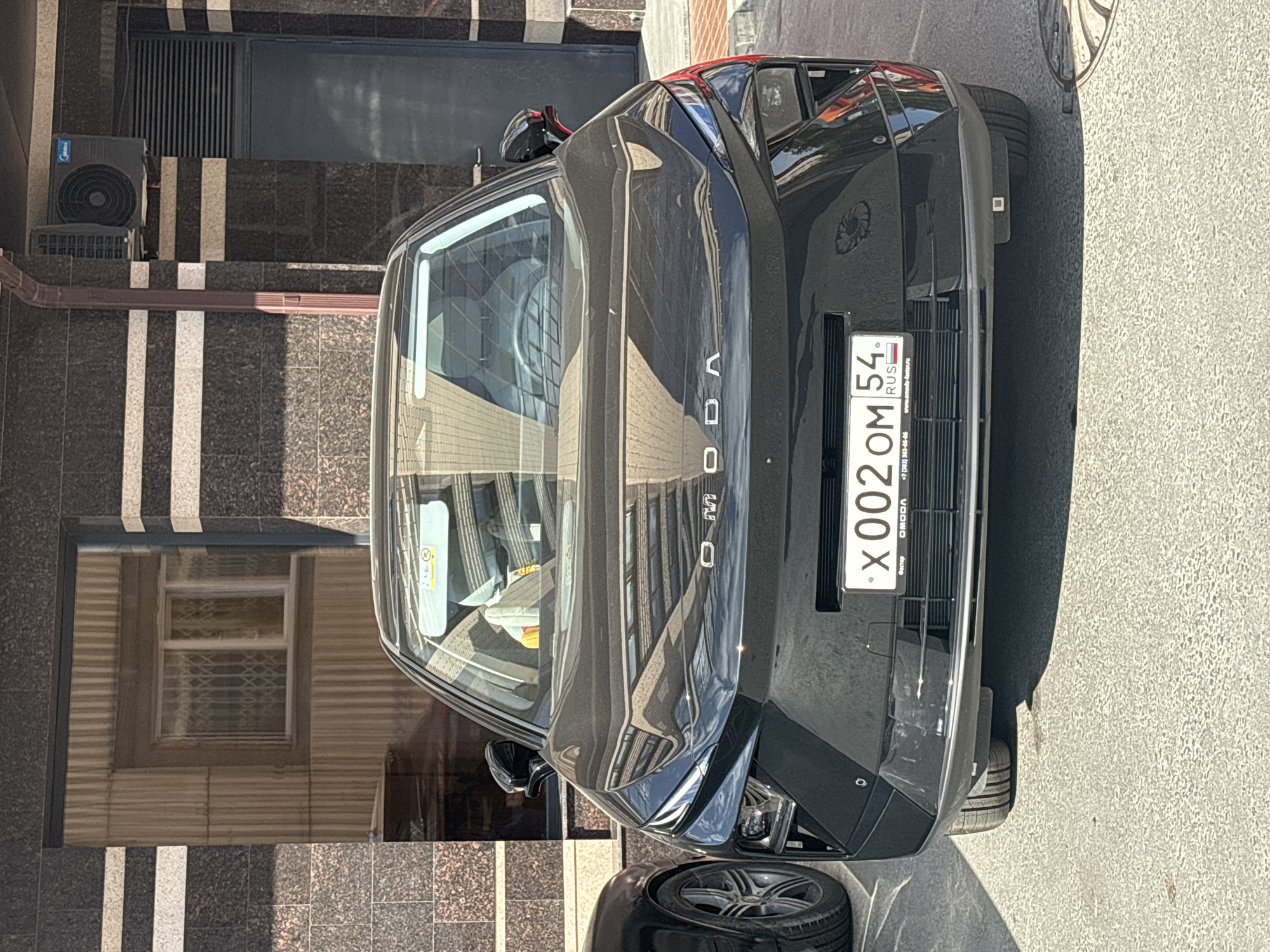 A black OMODA sedan is parked in front of a building in Novosibirsk, Russia. The car has a Russian license plate reading "X 002 OM 54". The building has a dark facade with horizontal light-colored stripes and a large window on the left side of the image, reflecting the sky and trees. An air conditioning unit is visible mounted on the building's exterior to the right of the window. The car's front is angled slightly towards the viewer, and its headlights and taillights are visible. The street in front of the car is paved and appears to be in good condition. The sun is shining, casting shadows on the ground.