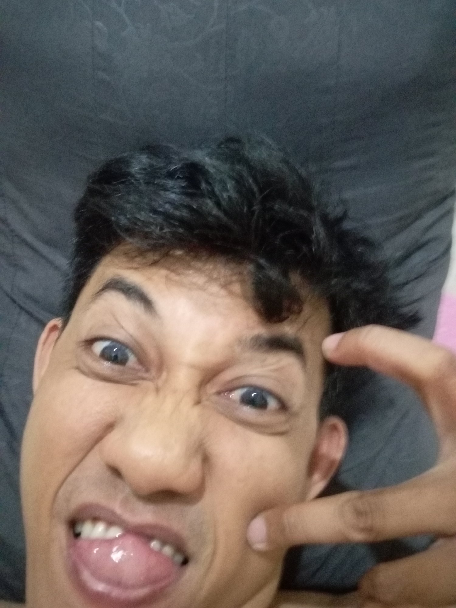 A close-up, low-angle selfie shows a person's face with their mouth open and tongue sticking out. Their eyes are wide and their eyebrows are furrowed, suggesting a playful or exaggerated expression. A finger is touching their cheek. The background is a dark, textured surface, possibly a bed or pillow, and a hint of pink fabric is visible on the right side. The lighting is dim and appears to be from above, casting shadows on the face. The image was taken in Batam, Indonesia.