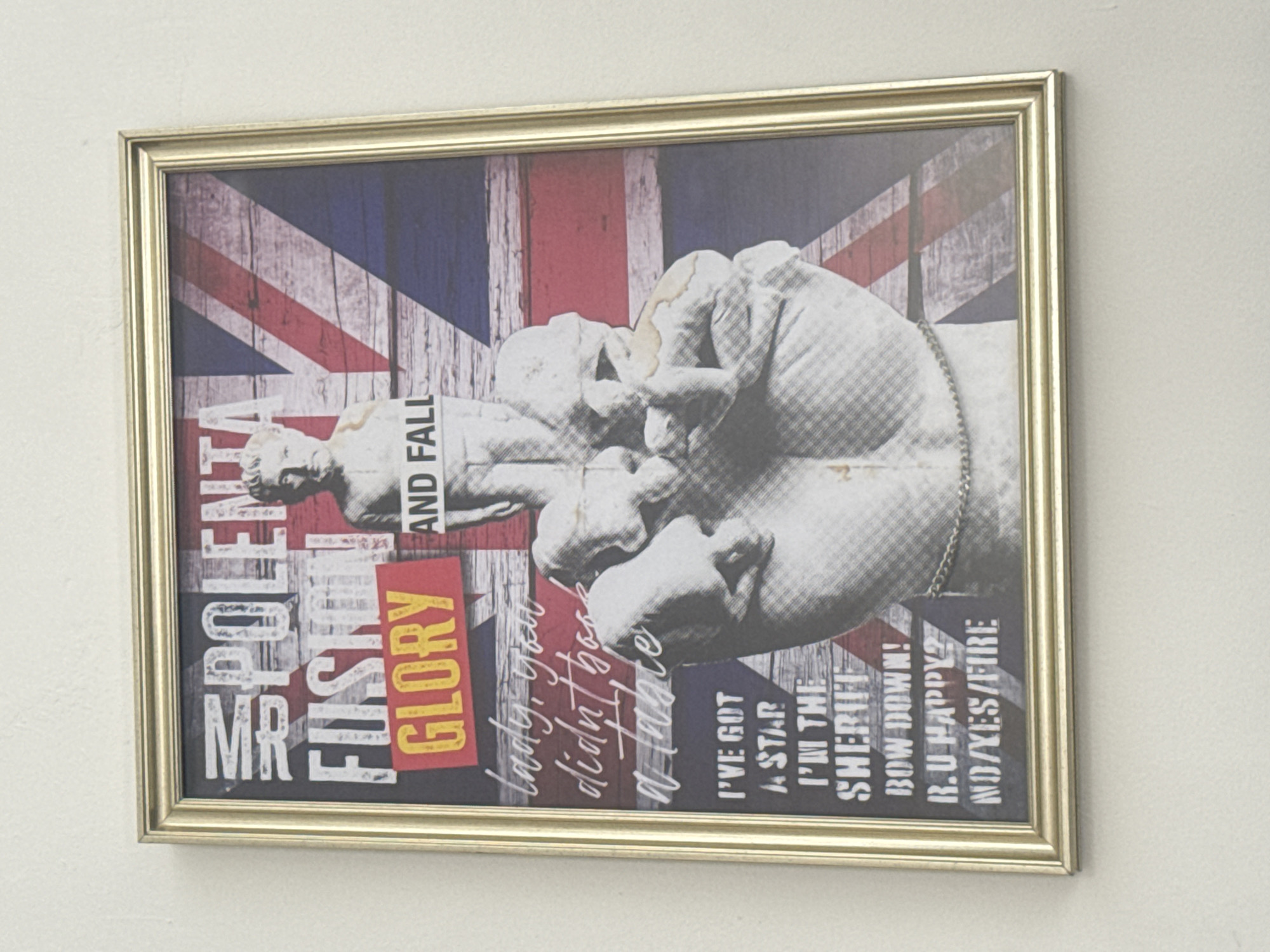 A framed print hangs on a white wall. The print features a British flag backdrop overlaid with a black and white image of a clenched fist wearing a chain, seemingly made of stone. Text in various styles and colors is scattered around the image, including "MR FIRST SPOLEMA", "FUSION GLORY", "AND FALL", "lady, you didn't book a Table", "I'VE GOT A STAR", "I'M THE SHERIFF", "BOW DOWN!", and "R.U HAPPY? NO/YES/FIRE". The frame is gold. The image is not identifiable as being located in Tel-Aviv, Israel.