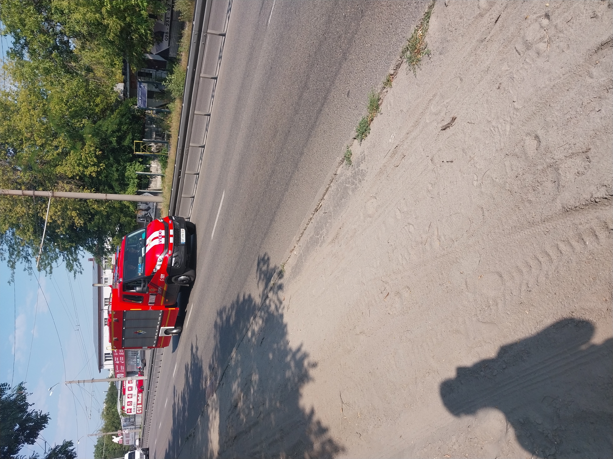 The image shows a red fire truck driving on a wide road in Dnipro, Ukraine. The fire truck is a modern Iveco vehicle with the number "101" visible on its side, indicating it is an emergency vehicle. The truck is in motion, heading away from the viewer and towards the right side of the frame.
The road is paved and appears to be a multi-lane highway or main thoroughfare. There is a metal guardrail separating the road from a sandy, unpaved area on the left side of the image. This sandy area is likely a sidewalk or verge, and it is partially covered by tire tracks, suggesting vehicle traffic.
In the background, there are trees lining the right side of the road, and some buildings are visible behind them. One building appears to be a retail or commercial establishment with signs in Ukrainian. The sky is clear and blue, indicating a sunny day.
A shadow of a person holding a phone or camera is cast on the sandy area in the foreground, indicating the photographer's presence. The overall scene depicts a common urban or suburban environment in Dnipro on a clear day, with an emergency vehicle in transit.