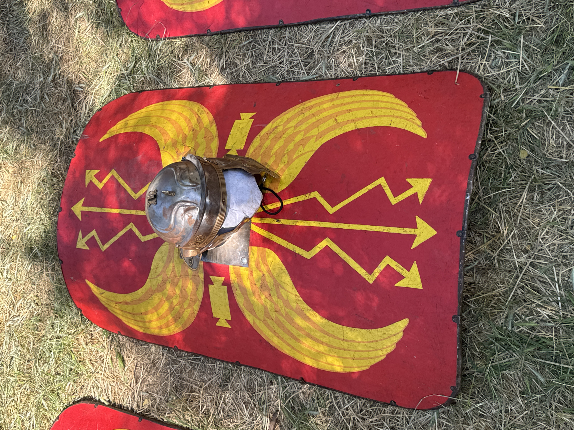 In the city of Plovdiv, Bulgaria, on a grassy area, a Roman legionary helmet rests on a red shield adorned with yellow designs. The shield features prominent wing-like motifs and lightning bolt patterns. The helmet is metallic and appears to be a replica, with visible rivets and detailing. Another shield, partially visible on the right, is also red with yellow markings. The ground is covered in dry grass and some green blades, suggesting an outdoor setting, possibly a historical reenactment or display. The lighting indicates it is daytime, with shadows cast by the surrounding environment.