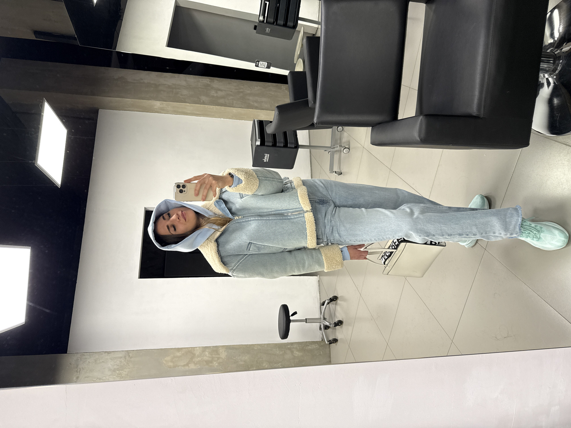 A woman is taking a selfie in what appears to be a hair salon. The woman is wearing a light blue hooded sweatshirt under a denim jacket with a cream-colored sherpa lining. She also wears light-wash jeans and light green sneakers. She is holding a white handbag in her left hand and a smartphone in her right hand, pointed towards her face.
The background of the image shows salon equipment, including black chairs on hydraulic stands, a small stool, and hair product dispensers. The walls are primarily white, with gray accents. The floor is light-colored with a geometric pattern. The lighting is bright and appears to be artificial.
The location is likely a hair salon in Saint Petersburg, Russia, given the typical salon decor and architectural style.
