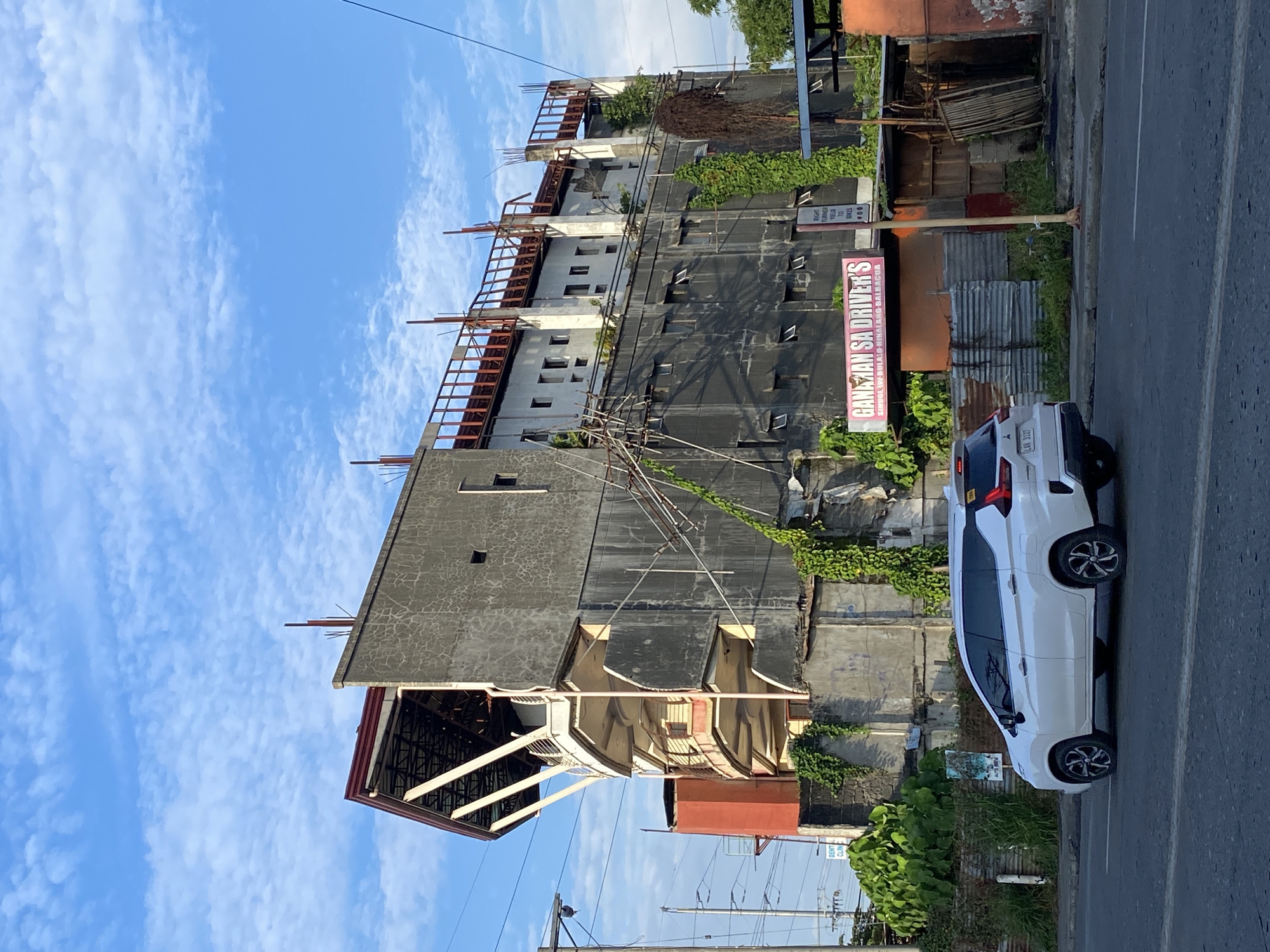 The image shows an unfinished building located along a street in Davao City, Philippines. A white multi-purpose vehicle is driving on the road in front of the building. The building appears to be undergoing construction, with exposed concrete and rebar visible. Various plants and vines are growing on and around the structure, suggesting a prolonged period of inactivity or neglect. A pink and white sign with text in an unfamiliar language is attached to the building, and a traffic sign is also visible. The sky is partly cloudy.