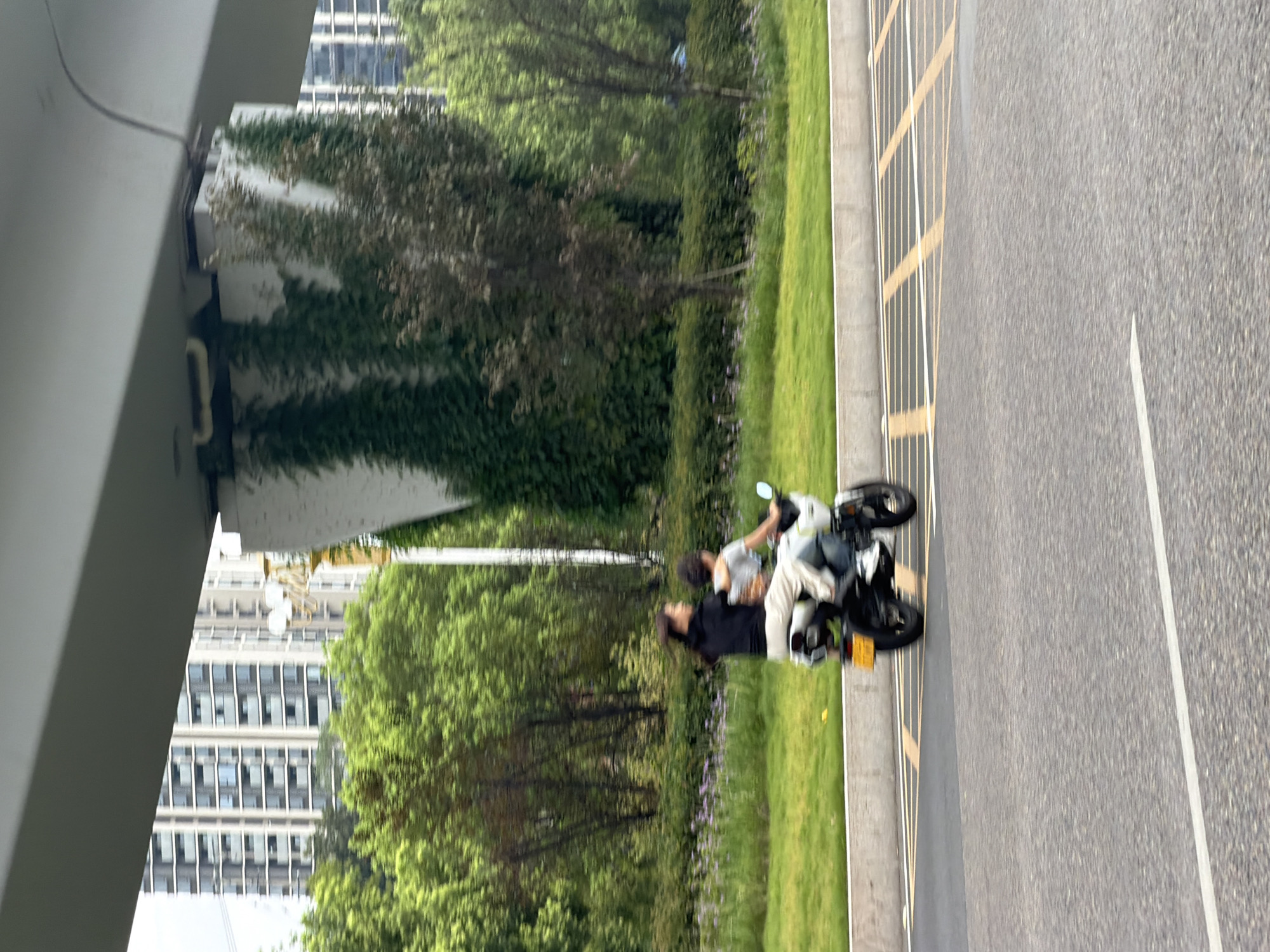 Two individuals are riding a motorcycle on a paved road in Jiangjin District, China. The person in front is wearing a black top and light-colored pants, and the person behind is wearing a gray top and blue jeans. The motorcycle is white. They are traveling under an elevated structure, possibly a bridge or overpass, which is supported by a concrete pillar covered in green ivy. In the background, there are lush green trees and tall modern buildings. The road has yellow and white lane markings. The scene appears to be during daylight.