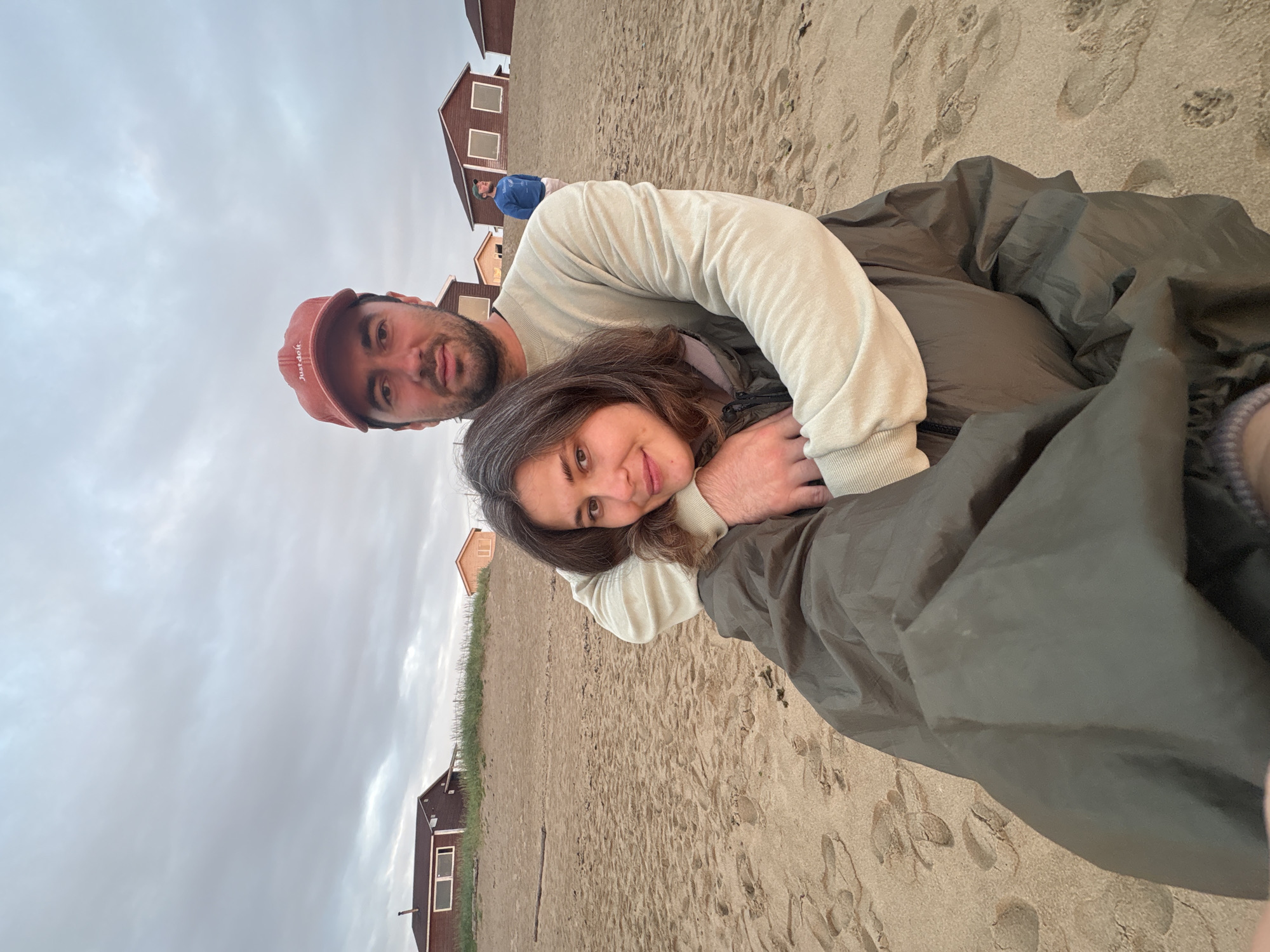 A medium shot captures a couple embracing on a sandy beach with a cloudy sky overhead, in Teriberka, Russia. The man, wearing a red baseball cap with the logo "Just do it." and a cream-colored sweatshirt, has his arms wrapped around the woman. The woman, with brown hair, leans into his embrace, resting her head on his arm. She is wearing a dark olive green jacket. In the background, several wooden houses with brown roofs are visible on a sandy incline, and another person in a blue jacket stands further up the beach. The sand is marked with footprints, suggesting recent activity. The overall atmosphere is serene, with the natural elements of sand, sky, and sparse structures dominating the scene.