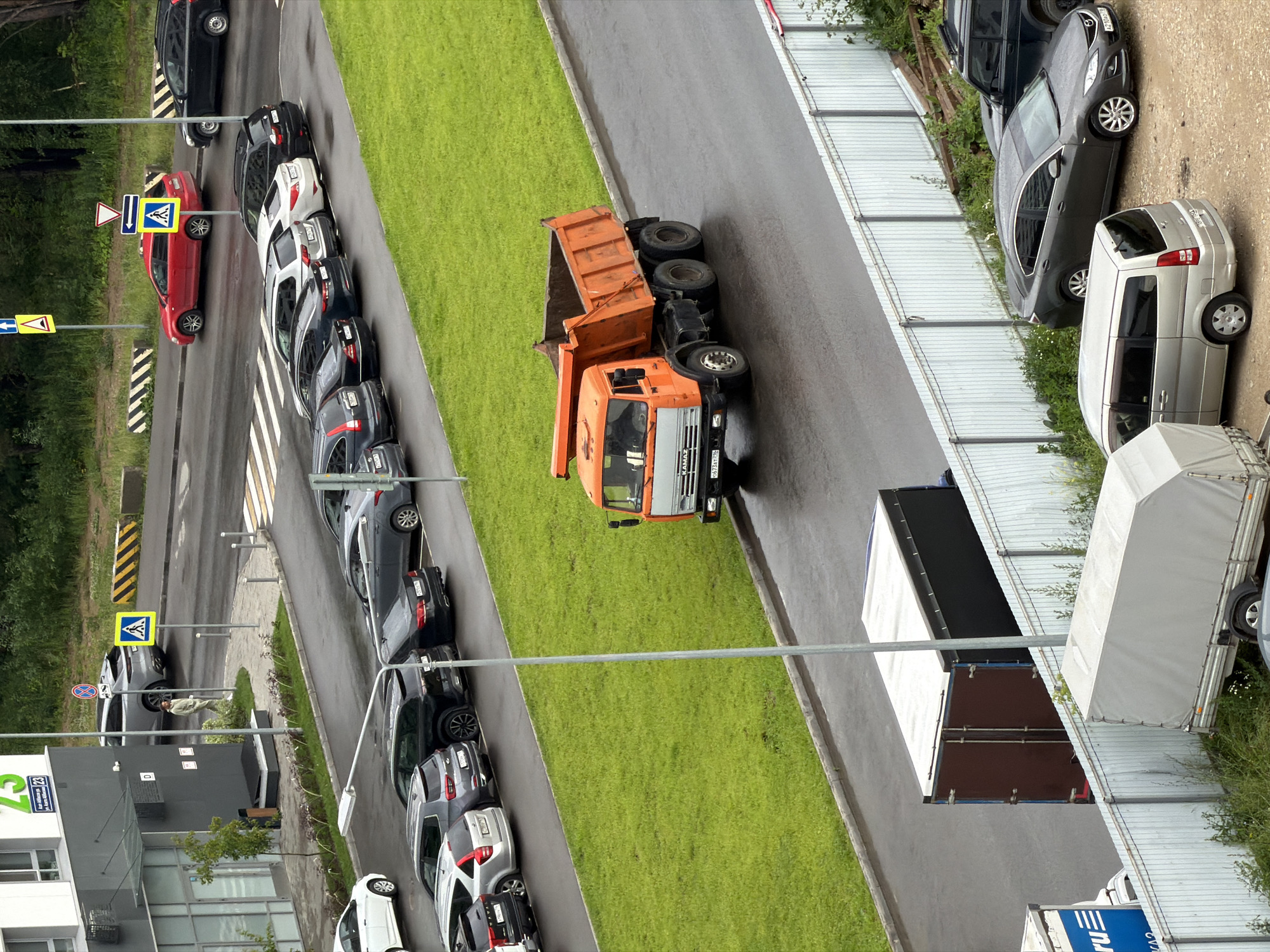 An aerial view captures an urban street scene with parked cars, a dump truck, and a building in Unknown, Unknown. A gray SUV is parked in front of a gray building with green signage. A person wearing a light-colored outfit stands on a sidewalk near a pedestrian crossing sign. The street is wet, suggesting recent rain, with several cars parked along its edge. Further down the street, an orange dump truck with dual rear wheels drives on the wet pavement. A white metal fence runs along the side of the street, separating it from a grassy area. Behind the fence, several trailers are parked, including a large dark-colored trailer with a white tarp, a silver minivan, and a dark gray sedan. A dark-colored SUV is parked in the background. The environment appears to be a commercial or residential area with trees and greenery visible in the distance.