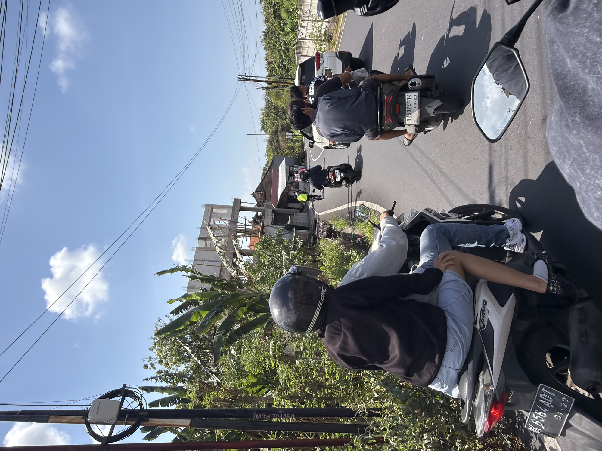 In Kuta Selatan, Indonesia, a sunny day unfolds on a road with lush greenery lining both sides. Several motorcycles are navigating the street. In the foreground, a motorcycle is seen from behind, with two individuals riding it. The passenger in the back, wearing a black helmet and jacket over a grey shirt, is seated with their legs crossed. The driver, partially obscured, is wearing a grey long-sleeved shirt. Their license plate reads "W 6564 ADI" with "01-27" below it.
Further down the road, another motorcycle with three passengers is moving away from the camera. The rider in front wears a dark shirt and shorts, while the two passengers behind him are also in dark clothing. Their license plate is visible as "DK 5024 FDK" with "08-29" below it.
A white SUV follows this motorcycle. In the distance, a person on a bicycle is seen on the left side of the road, and a construction site with unfinished concrete structures is visible behind the greenery. Telephone poles and wires crisscross the clear blue sky, dotted with white clouds.