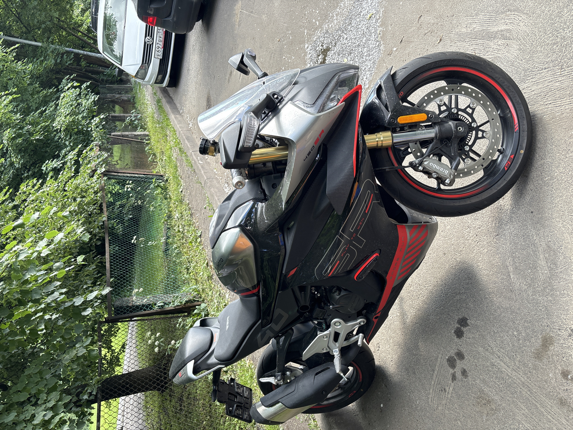 A black and silver sport motorcycle with red accents is parked on a paved surface next to a curb. The motorcycle has a clear windshield, a black seat, and a license plate. In the background, there is a chain-link fence, trees with green leaves, and a white Volkswagen car with a Russian license plate that reads "E697BY 11". Part of a dark-colored car is visible to the right of the motorcycle. The ground has some scattered debris and tire marks. The scene appears to be an outdoor urban environment.