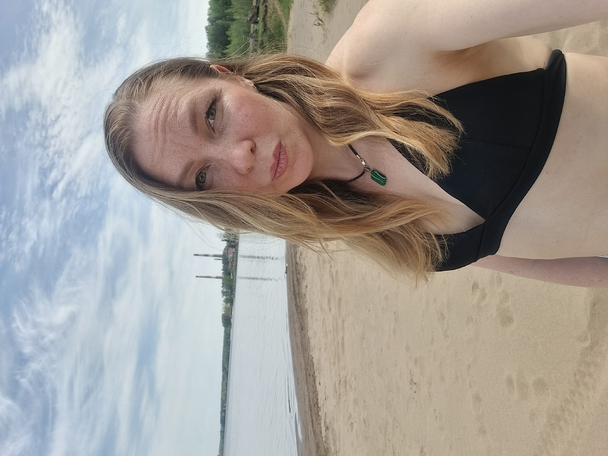 A woman with light brown hair and fair skin, wearing a black bikini top and a necklace with a green pendant, is positioned on a sandy beach. The location appears to be the city of Chaikovsky, Russia, as evidenced by the industrial chimneys visible in the background. The beach borders a body of water, possibly a river or lake, under a partly cloudy sky.
