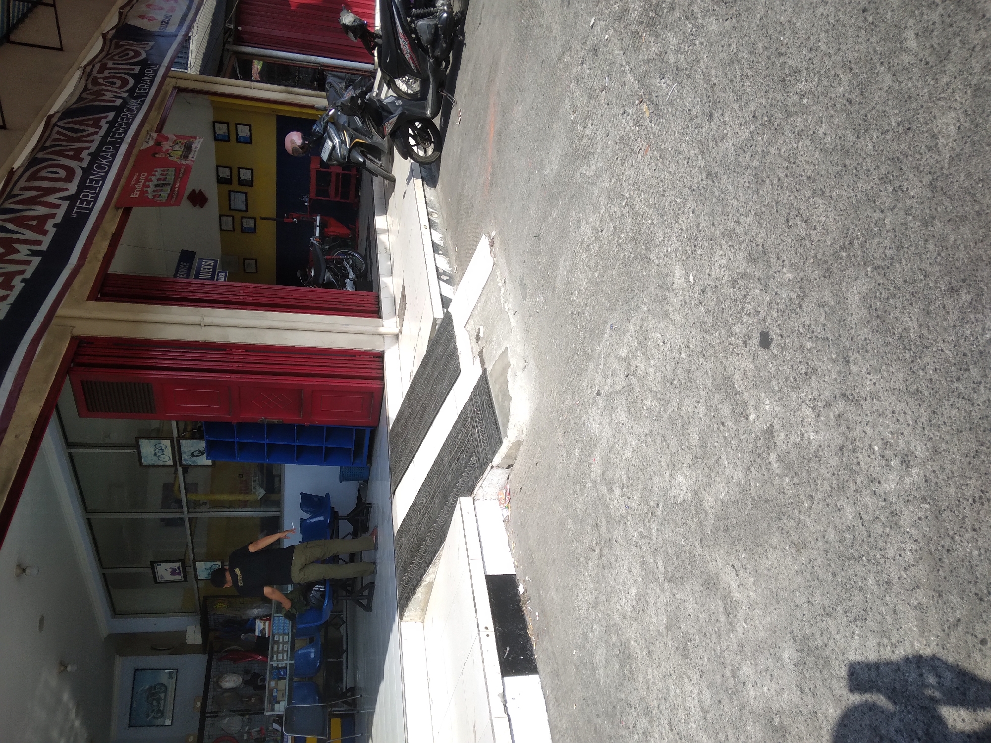 The image shows a view of "MANDAKA MOTOR", a motorcycle repair shop located in Purwokerto Kidul, Indonesia. A man wearing a black t-shirt and olive-green pants is standing near the entrance of the shop, holding a cigarette. The shop has red sliding doors and a banner with "MANDAKA MOTOR" printed on it. Inside the shop, several motorcycles are visible, along with tools and shelves stocked with parts. Two motorcycles are parked outside the shop. A ramp with a textured surface leads up to the shop's entrance. The ground in front of the shop is paved with asphalt. The environment appears to be a commercial area with other buildings visible in the background.