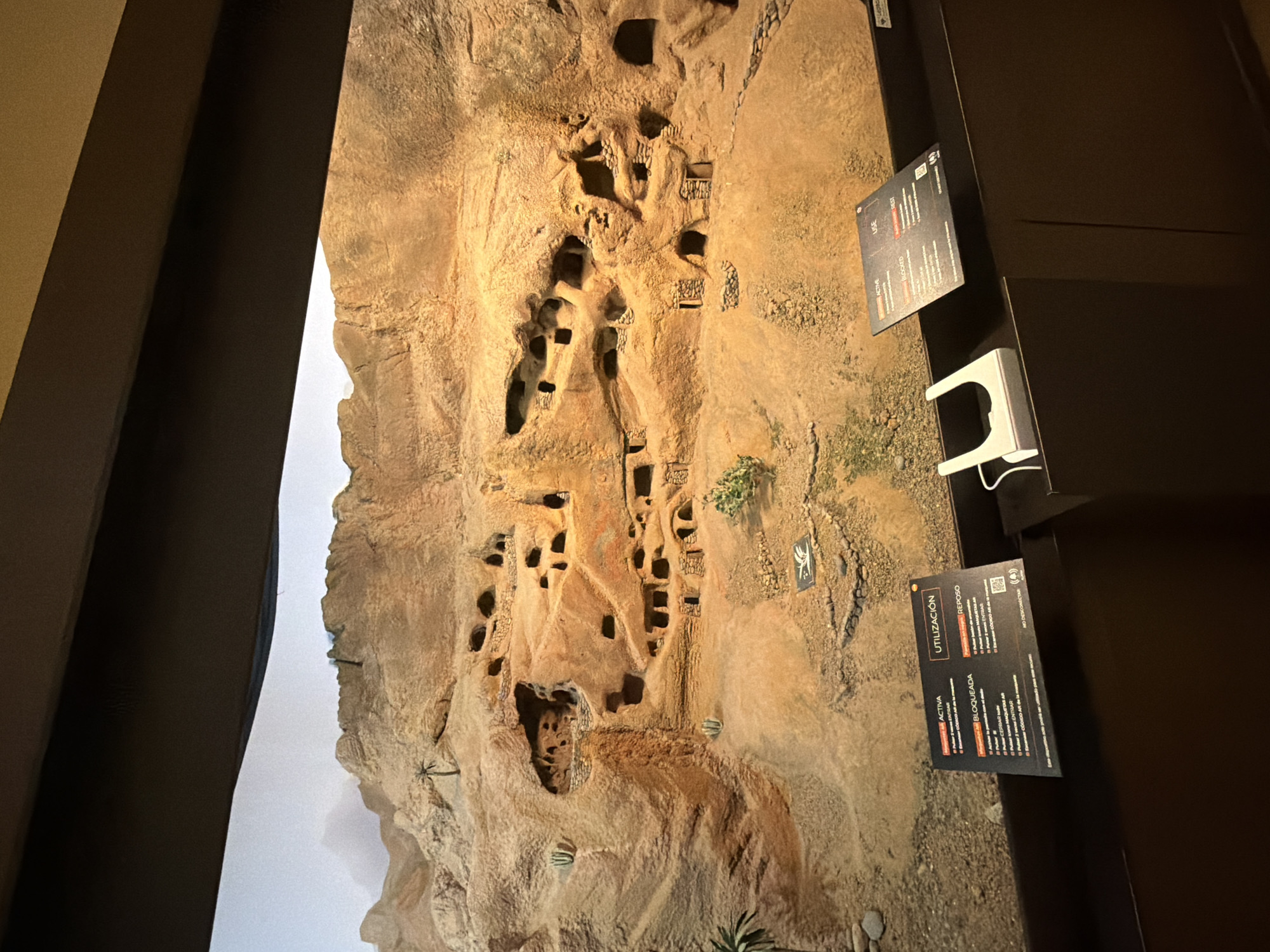 This is a medium shot, eye-level, indoor photograph of a diorama depicting a cliffside with numerous cave dwellings. The diorama is illuminated with warm lighting, highlighting the textured, sandy-brown rock formations. The caves, varying in size and shape, are carved into the cliff face, with some showing indications of stone structures around their entrances. Small, sparse vegetation, including what appear to be cacti and a palm tree, are scattered throughout the diorama, suggesting a dry, arid environment.
In the foreground, resting on a dark brown shelf, are two informational display cards and a white device. The card on the left, partially obscured by the white device, has text in Spanish. The card on the right, also in Spanish, has sections labeled "UTILIZACIÓN" (Utilization) and "REPOSO" (Rest), and appears to provide instructions for operating the white device. The white device is a holder for what is likely a smartphone or tablet, with a cable attached.
The background of the diorama shows a more distant, hazy mountain range under a pale sky. The overall scene is a detailed model representing an ancient or historical settlement built into a rocky landscape. The context of Las Palmas De Gran Canaria, Spain, suggests this diorama is likely found in a museum or cultural exhibition within that city, potentially depicting a historical site or archaeological find relevant to the Canary Islands. The presence of the informational cards and the device indicate an interactive exhibit. No people are visible in the image.