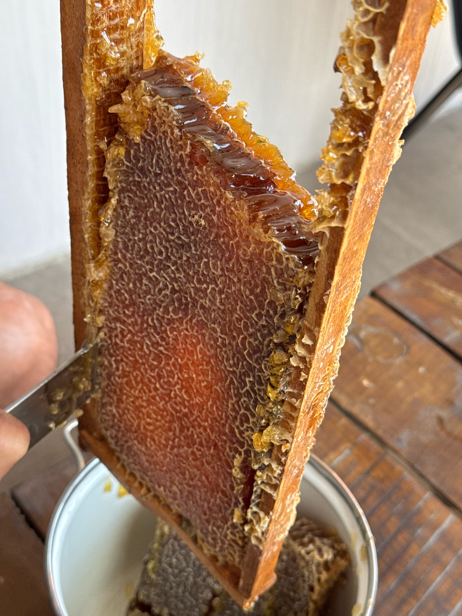 A close-up, top-down shot shows a person using a knife to cut a piece of honeycomb from a wooden frame. The honeycomb is a deep amber color and appears to be filled with honey. The frame is suspended over a white bowl, with some honeycomb already collected inside. The background consists of a wooden table and a white wall. The scene is likely taking place in Telavi, Georgia, a region known for its beekeeping traditions.