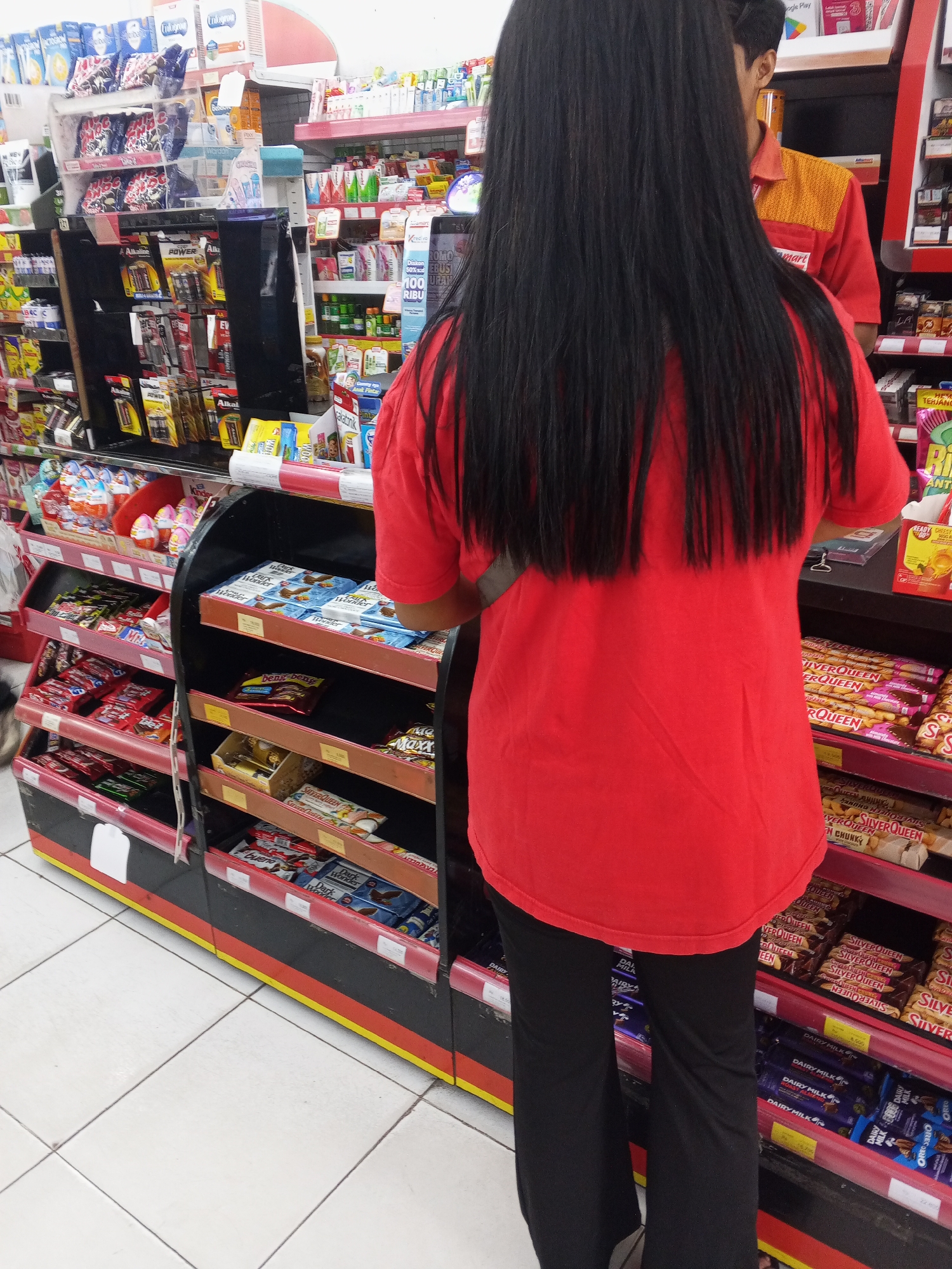 A woman with long black hair, wearing a red t-shirt and black pants, stands in an aisle of a convenience store. She is facing away from the camera, looking at shelves stocked with various products, including candy bars, batteries, and personal care items. To the right of the woman, a male store employee in a red and yellow uniform is partially visible. The store appears to be a Minimart or Indomaret, common convenience store chains in Indonesia. The tiled floor and brightly lit shelves are typical of such establishments. The scene is likely captured in Ciruas, Indonesia, given the presence of these store types.