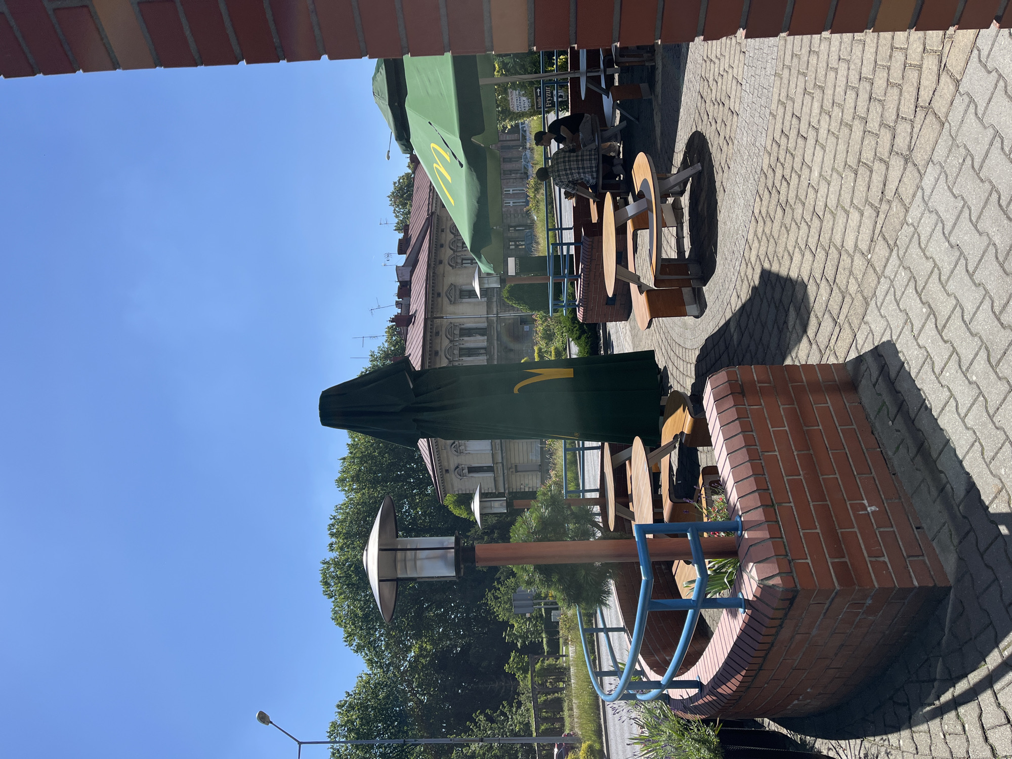 The image shows an outdoor seating area for a McDonald's restaurant on a sunny day. Two people are seated at a round table under a green McDonald's umbrella. The tables and chairs are made of wood and are arranged on a paved patio. In the background, there are buildings and trees. A street lamp is visible on the left side of the image. The sky is clear and blue, indicating good weather. The lighting suggests it is midday. The scene conveys a relaxed atmosphere, with patrons enjoying the outdoor dining experience. There is some visible text on the green umbrella which appears to be the McDonald's logo.