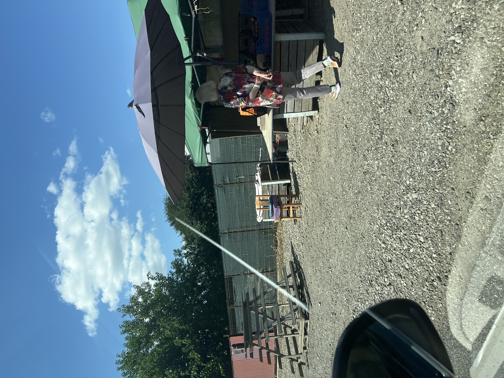 In Rădășeni, Romania, on a bright, sunny day with scattered clouds, an outdoor market stall is set up. A woman with short blonde hair, wearing a colorful floral-patterned shirt and grey pants, stands near a table laden with produce under a large gray umbrella and a green canopy. She is facing away from the camera with her hands clasped behind her back. In the foreground, a car's side mirror and the edge of its windshield are visible, indicating the photo was taken from inside a vehicle. The ground is covered in gravel. To the left, a rustic wooden tiered structure, possibly for display or seating, stands near a corrugated metal fence. A single wooden chair with colorful cushions sits to the right of the structure. The environment suggests a rural or semi-rural setting, likely a local market or farm stand.