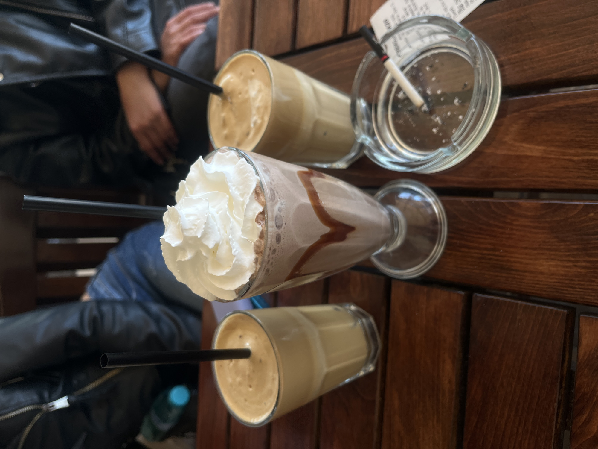 The scene depicts a wooden table in an outdoor setting. Three drinks are arranged on the table: two glasses filled with a tan-colored beverage with black straws, and a tall glass containing a chocolate-colored drink topped with white whipped cream. An ashtray with a lit cigarette is positioned near one of the tan drinks. Two people are partially visible around the table, wearing dark-colored jackets. A water bottle is visible under the table. The location is Karnobat, Bulgaria.