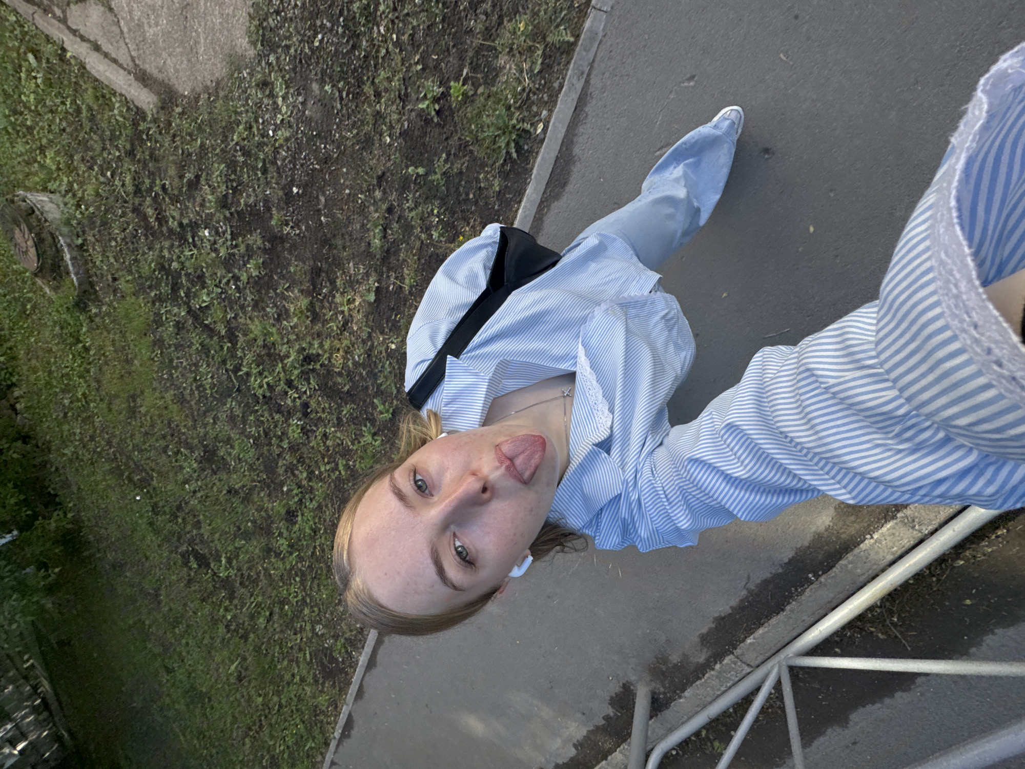 A top-down, angled outdoor shot shows a young, light-skinned person with blonde hair sticking out their tongue. The person wears a light blue and white striped long-sleeved shirt, light blue jeans, and white sneakers. A black bag strap is visible across their chest. The location appears to be near a sidewalk or paved area adjacent to a grassy area with some sparse ground cover. A metal railing is visible in the lower right corner. The setting suggests an urban environment. While the specific location in Perm, Russia, cannot be definitively confirmed from the image, the style of buildings and foliage are not inconsistent with that region.