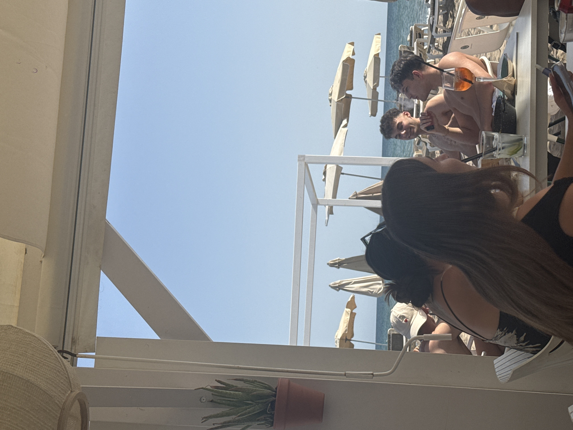 This image depicts a group of people seated at a table on a sunny day at a beachside establishment in Castelldefels, Spain. In the foreground, a person with dark hair is partially visible, facing away from the camera and holding a mobile phone. To their right, a man with short, dark curly hair is smoking and looking to his left, holding a cigarette. Next to him, another man with short, dark curly hair is looking to his right, smiling and resting his chin on his hand. A glass of orange liquid with a straw, possibly Aperol Spritz, sits on the table in front of the man who is smoking. A glass with ice and a lime, along with a black bowl, are also on the table. In the background, beach umbrellas and lounge chairs are visible on a sandy beach, with the blue Mediterranean Sea and clear sky beyond. A potted plant hangs on a white post in the left foreground. The overall atmosphere is casual and relaxed, suggesting a vacation or leisure setting.