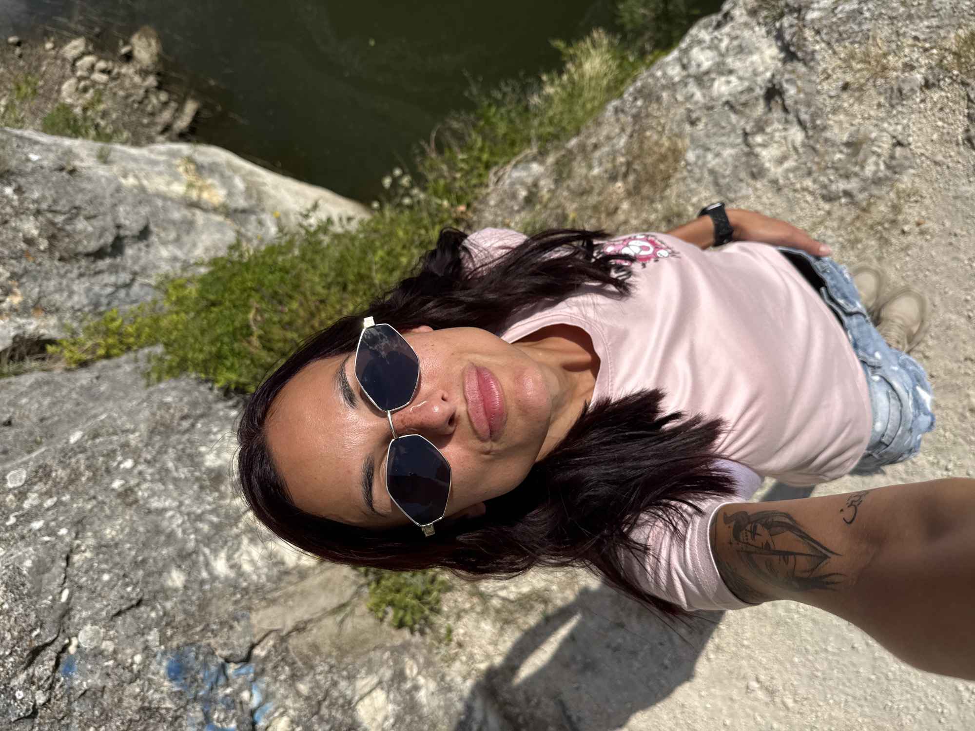 A woman is taking a selfie while standing on a rocky cliff overlooking a body of water. She has dark hair, is wearing sunglasses, a pink t-shirt, and denim shorts. Her left arm, which is extended to take the photo, is adorned with a tattoo. The background shows a rocky cliff face with some greenery and a dark, reflective body of water. This location is likely near the Kaylaka Park in Pleven, Bulgaria, known for its scenic cliffs and lake.