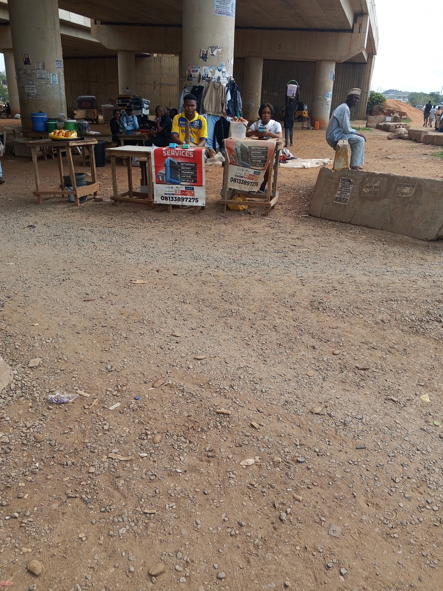 The image depicts a street scene in Galadimawa Village, Nigeria, under an overpass. The ground is dirt and gravel. Several individuals are present. A man in a yellow and blue shirt sits at a table with a sign advertising financial services like withdrawals, deposits, transfers, airtime, and bill payments. A woman in a white shirt sits at a similar table with a similar sign. An elderly man in a patterned blue outfit sits on a concrete barrier. Other people are milling around in the background, near the tables. Behind the tables, additional people are seated around another table. There are various makeshift tables, containers with food, and clothes hanging under the overpass. The overpass itself has pillars covered in posters and advertisements.