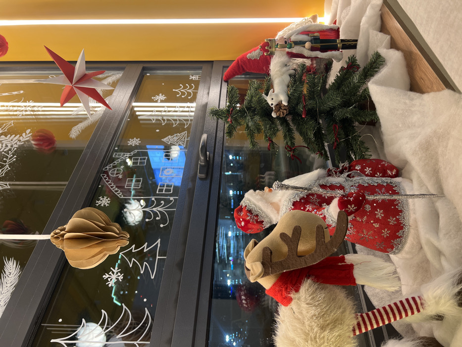 The image depicts a detailed Christmas-themed window display located in Krasnoyarsk, Russia. In the foreground, a layer of white, fibrous material simulates snow on a wooden shelf. From left to right on this "snow," a collection of holiday figures is arranged: a tall, light-brown reindeer figure with felt antlers and a red scarf; a Santa Claus or Ded Moroz figure in a red, silver-embroidered coat and hat, holding a staff and a red sack; a small artificial green Christmas tree adorned with red bows and a white squirrel ornament holding a pinecone; and a partially visible Nutcracker figure in a red and blue uniform with a gold crown.
Behind the display, a large window features white decals of Christmas trees, snowflakes, and gingerbread houses. Hanging from the top window frame are a folded brown paper ornament and a red and white paper star. The window reflects interior yellow lighting and, beyond, presents a blurry, out-of-focus nighttime urban scene with discernible streetlights and distant building lights. To the far right, a vertical section of a yellow wall with illuminated vertical light strips is visible.