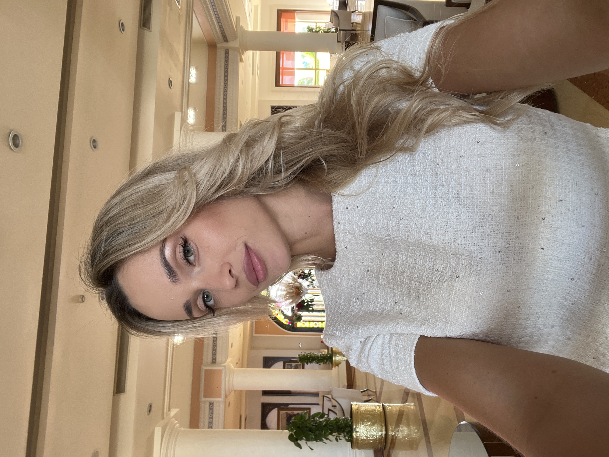 A young, fair-skinned woman with long, wavy blonde hair is taking a selfie in a well-lit, indoor hotel lobby. She is wearing a textured white, short-sleeved dress with subtle sequin embellishments. Her makeup includes winged eyeliner and pink lipstick. The background features ornate pillars, reflective flooring, large potted plants, and arched doorways with colorful stained glass. The architecture suggests a luxurious resort, potentially located in Sharm El Sheikh, Egypt, known for its elaborate hotel designs. The woman is positioned centrally, facing the camera with a soft smile.
