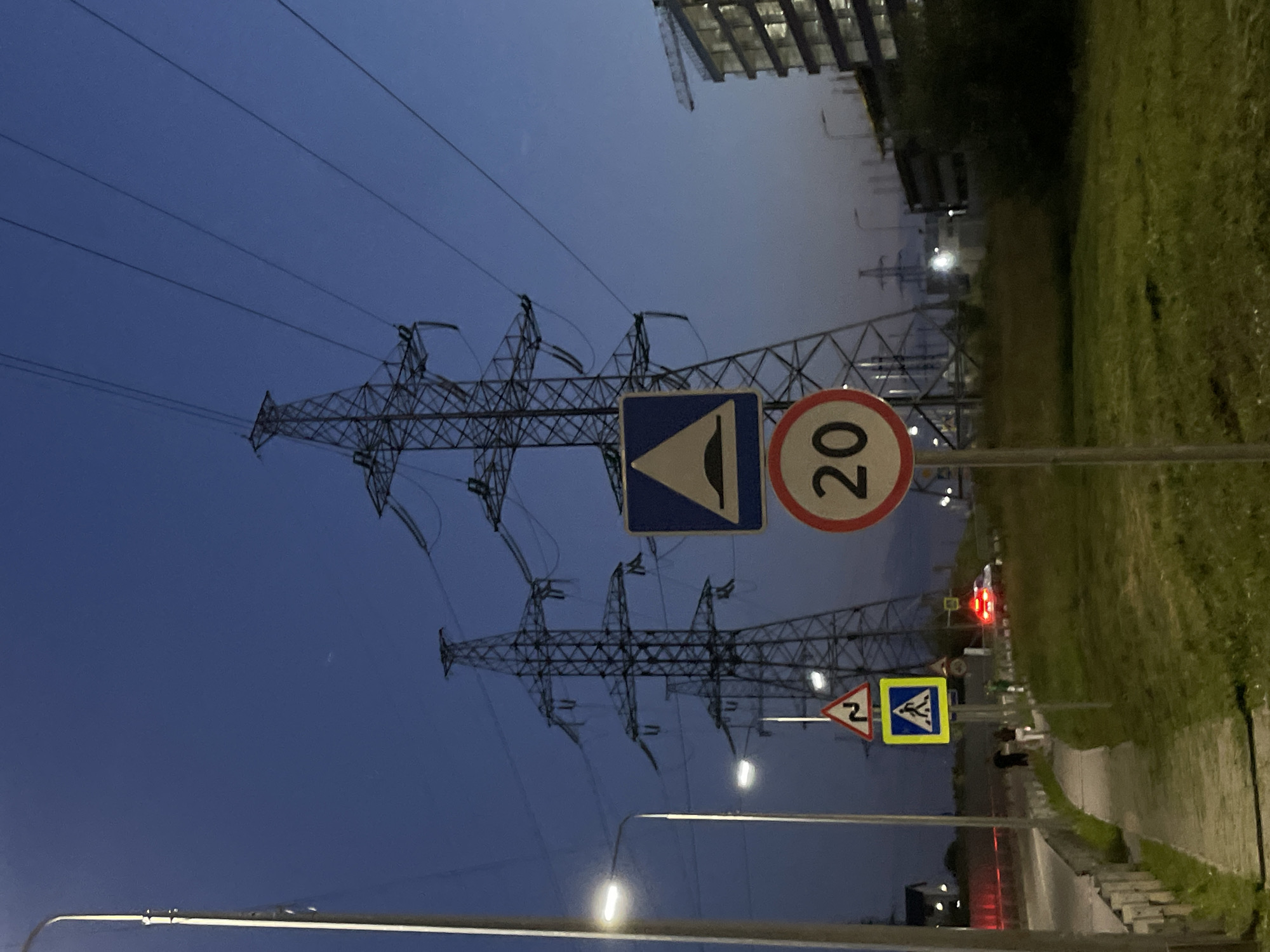 The image depicts a roadside scene at dusk in Odintsovo, Russia, with multiple power line towers dominating the background. A paved walkway runs along the side of a road, separated by a low concrete barrier. Several road signs are visible: a blue and yellow pedestrian crossing sign, a red-bordered sign with a white triangle depicting a bumpy road, and a circular speed limit sign showing "20". Two streetlights illuminate the walkway. In the middle ground, a white car with red taillights is stopped or moving slowly on the road. A few pedestrians are visible on the walkway. Further in the background, a multi-story building under construction is partially illuminated, with a construction crane visible. The sky is a deep blue, indicating twilight.