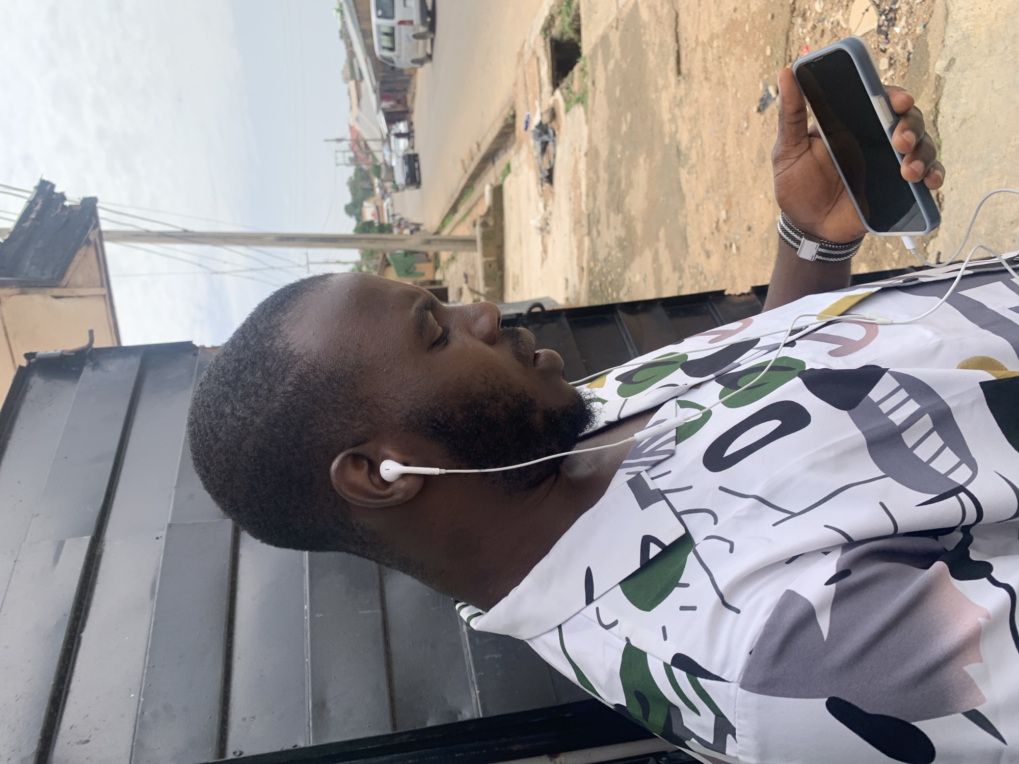 The image shows a close-up, vertically oriented shot of a Black man wearing earbuds and a white shirt with a bold, abstract pattern of green, gray, and black shapes. He is holding a smartphone with a light gray case in his right hand, which is positioned in the lower right quadrant of the frame. A white wire connects the earbuds to an unseen device. The man has a short haircut and a beard. His eyes are closed, and his head is tilted back slightly, suggesting he is relaxed or listening to something.
The background is blurred, but it appears to be an outdoor setting with a street and buildings. Hints of the environment include a paved area, some foliage, and what looks like a dark metal structure in the foreground. The lighting suggests it is daytime, possibly overcast, given the diffused light and the absence of strong shadows.
The man's attire, the presence of the smartphone, and the earbuds suggest a moment of leisure or engagement with technology. The overall mood is calm and personal. The location context of Jos, Nigeria, combined with the visible environmental elements, points to an urban or semi-urban street scene. There is no visible text in the image.