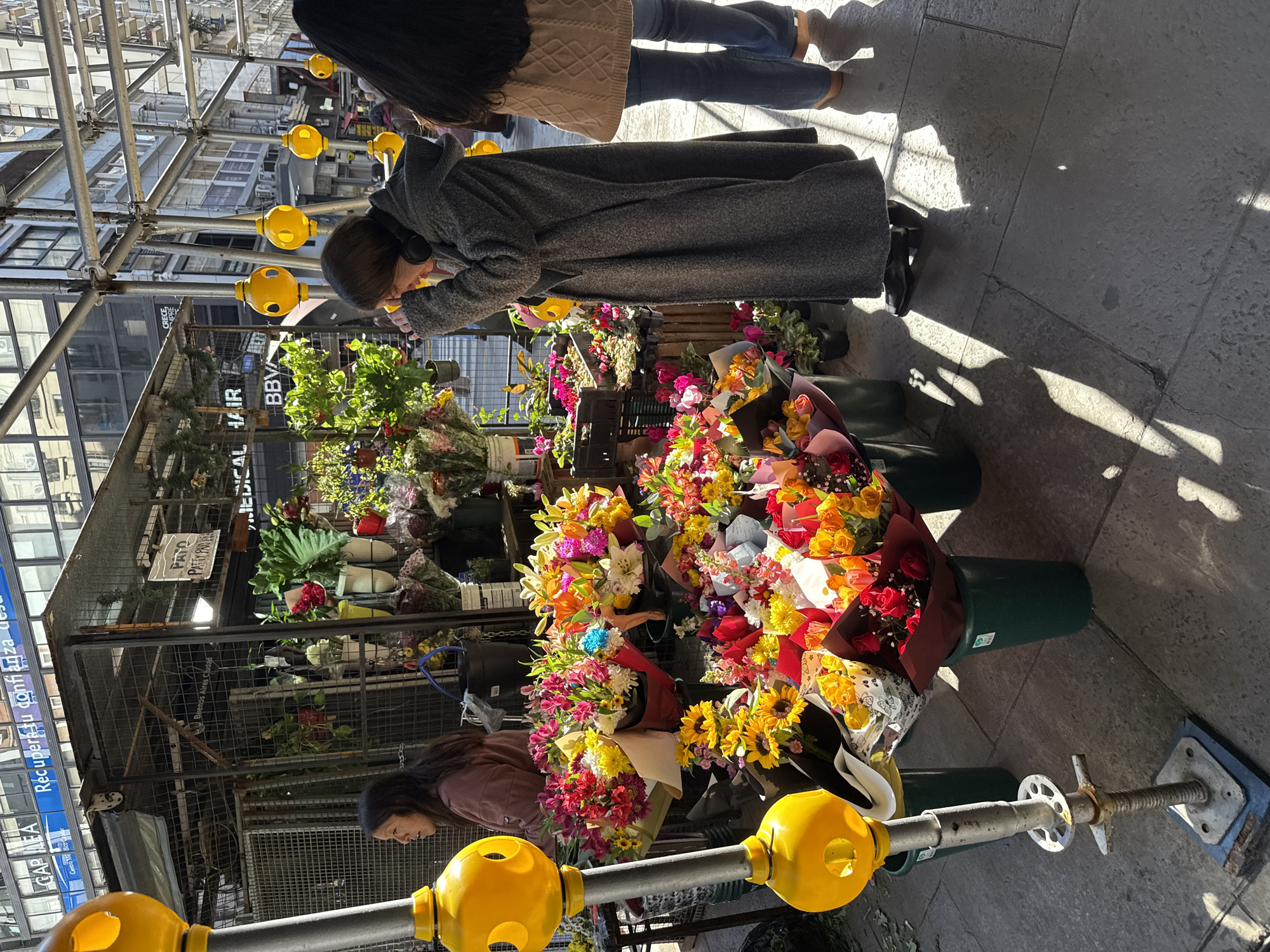 The photograph depicts a flower stand on a city street, likely in Buenos Aires, Argentina, as street signs show that city’s name. A woman in a long grey coat is browsing the flowers. Another woman in a brown jacket is lower down, also looking at the flowers. The flowers are arranged in bouquets and displayed in green buckets. The scene is framed by construction scaffolding with yellow spherical safety caps. Signage indicates “MEDICAL HAIR” and “BBVA” are nearby, along with “CRECE” and “PATO PATTYPAD.” The street surface is paved with large stone tiles. Several other people are visible in the background.