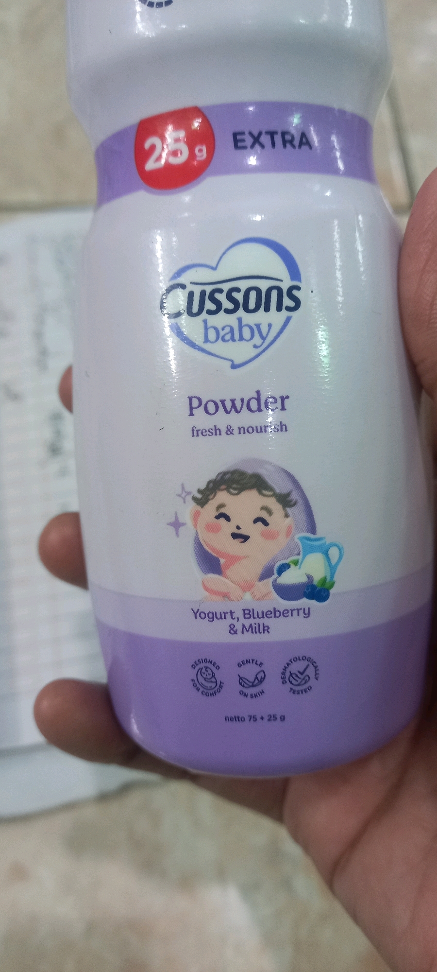 The image shows a close-up of a Cussons baby powder bottle. The bottle is primarily white with purple accents. On the front of the bottle, there is a red circular label with "25 g" printed in white, and next to it, the word "EXTRA" in white. Below this, the brand name "CUSSONS baby" is displayed in a blue heart shape. Underneath the brand name, the word "Powder" is written in purple, followed by "fresh & nourish" in smaller text. A cartoon illustration of a smiling baby with a jug of milk and blueberries is also visible. At the bottom of the bottle, there are three circular icons with text indicating "DESIGNED FOR COMFORT", "GENTLE ON SKIN", and "DERMATOLOGICALLY TESTED". Below these icons, the net weight is indicated as "netto 75 + 25 g". The bottle is held by a person's hand, with fingers visible on the left and right sides of the image. The background is out of focus and appears to be a wall or countertop, with some faint lines suggesting a patterned surface. The scene is indoors, and the lighting suggests a typical indoor environment. There is no visible information in the image that places the scene specifically in Sedati Agung, Indonesia.