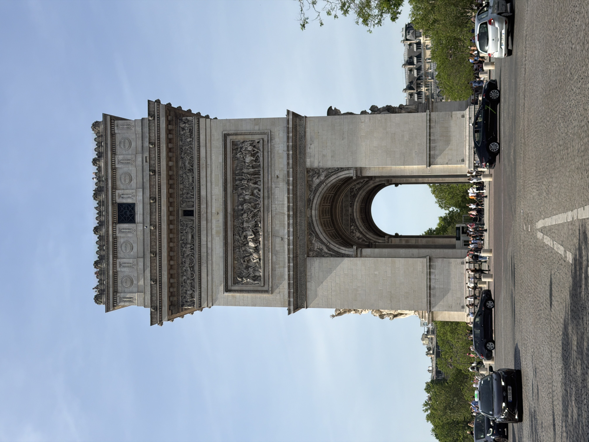 The image captures the Arc de Triomphe in Paris, France. The architectural structure is centrally positioned, showcasing its detailed carvings and arches. On the right, a cobblestone street is visible with several cars parked alongside it. A crowd of people lines the street, observing the monument. The sky is clear and bright, suggesting daylight conditions.