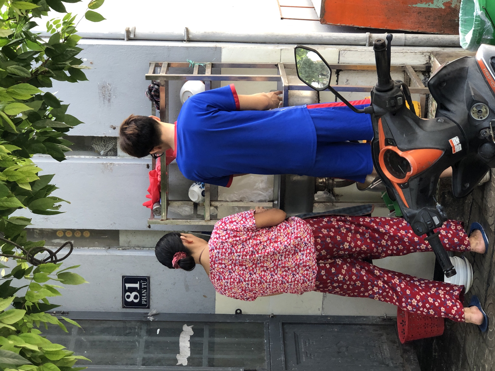 A two-person scene takes place outdoors on a tiled ground. The background shows a pale grey wall with a sign indicating "81 PHAN TƯ", a dark metal door to the left, and a metal shelving unit to the right with various items. A scooter is parked in the foreground.
On the left, a woman with black hair tied in a bun with a red scrunchie is facing away from the camera. She is wearing a red floral patterned short-sleeved shirt and matching red floral patterned pants, with blue flip-flops on her feet. She stands next to a red plastic basket.
To the right of the woman, a person with short brown hair, wearing glasses, is also facing away from the camera. They are dressed in a bright blue t-shirt with red trim and blue shorts with red trim. They appear to be interacting with the shelving unit, which holds white bowls and other kitchenware, some covered in plastic.
The overall environment suggests a residential or commercial area in Đà Nẵng, Vietnam, possibly near a street or alleyway. The lighting indicates it is daytime, with sunlight filtering through overhead green leaves.