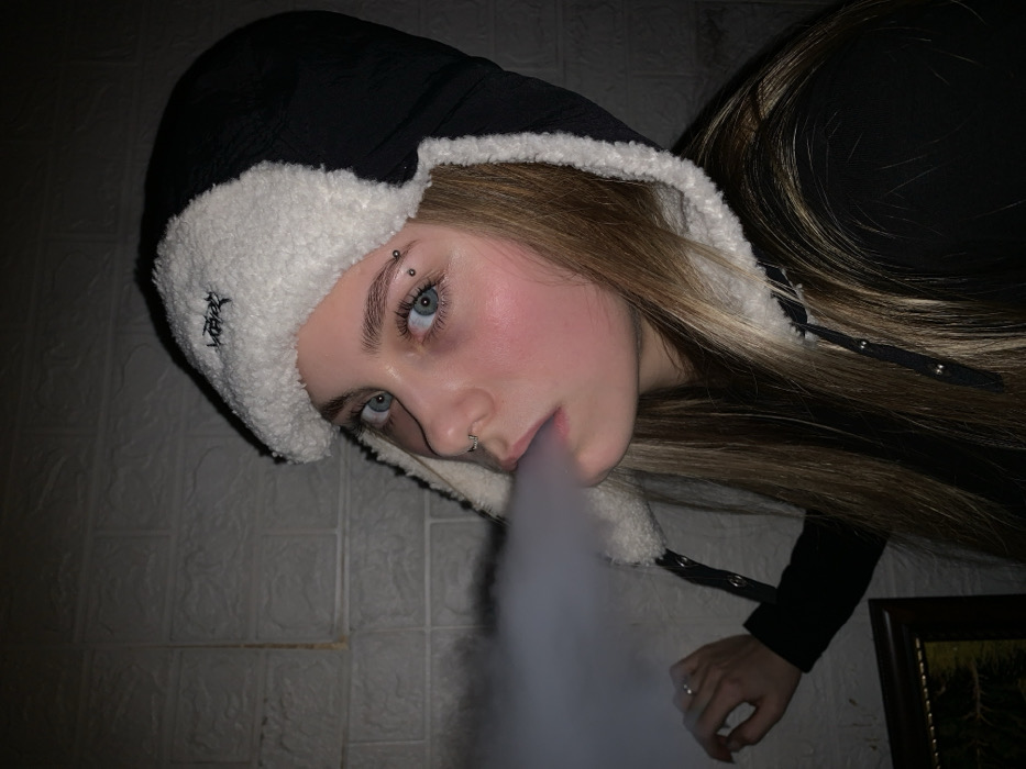 The image features a young person, likely female, exhaling a cloud of vapor or smoke. The person's head is tilted significantly to the left, giving the image a rotated appearance. They have fair skin, striking light blue eyes, and long, straight brown hair with lighter, possibly blonde, streaks that flow over their right shoulder and extend to the bottom right of the frame.
The individual is wearing a distinctive black and white hooded hat, possibly a trapper hat or similar style, made of a black fabric exterior and a plush, furry white interior lining that frames their face and extends around the ears. On the white furry trim near the left ear, there is black embroidery, which appears to be a stylized logo or text, though it is not clearly legible. Black straps with silver snaps are visible on the hat, one near the chin area. The person is also wearing a dark-colored top or jacket.
Notable facial features include an eyebrow piercing above their left eye (viewer's right), consisting of a silver bar with two small silver balls. They also have a silver septum piercing (a small hoop) in their nose. Their cheeks show a rosy flush, and their gaze is directed upwards and slightly to the right, creating an intense expression.
The primary activity depicted is the exhalation of a thick, white, voluminous cloud of vapor or smoke from their mouth, which streams downwards towards the bottom center of the frame.
The background is dimly lit and appears to be an indoor setting. To the left, a wall with a textured pattern resembling bricks or tiles is visible in a muted, possibly off-white or light grey, shade. To the lower right, a small portion of a dark-framed object, possibly a picture or mirror, is visible. A hand, likely the person's, is partially visible in the lower right corner of the frame, wearing a simple ring on one finger. The overall lighting is low-key, with the person's face and the vapor cloud being the brightest elements.