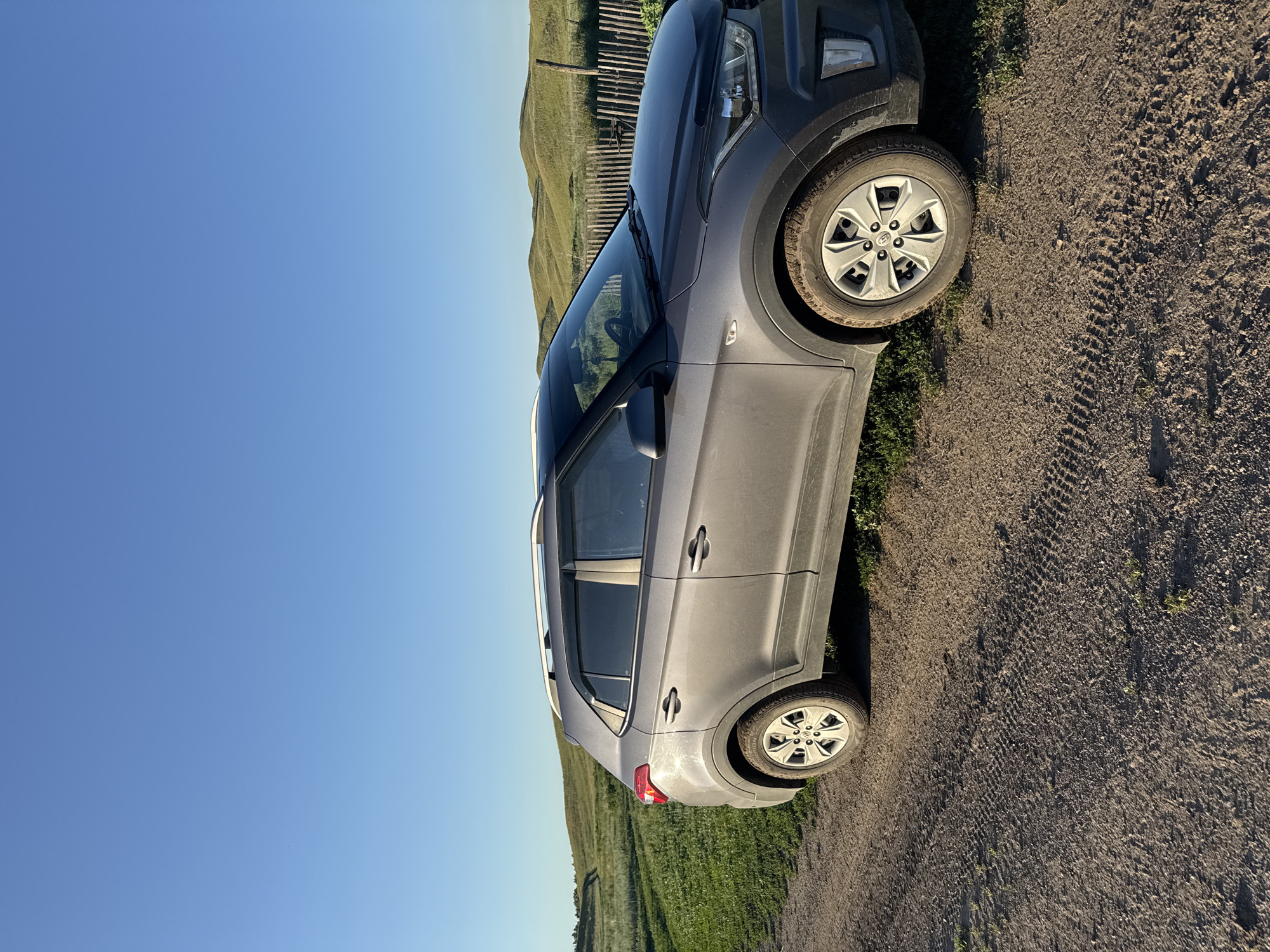A grey SUV is parked on a dirt road in Starokhalilovo, Russia. The car is viewed from the side, with its front right tire on the road and its rear right tire on grass. The car has tinted windows and a roof rack. In the background, there is a wooden fence and rolling green hills. The sky is clear and blue, indicating a sunny day. The image appears to have been taken in the late afternoon or early evening, as suggested by the warm lighting and long shadows. There is no text visible in the image.