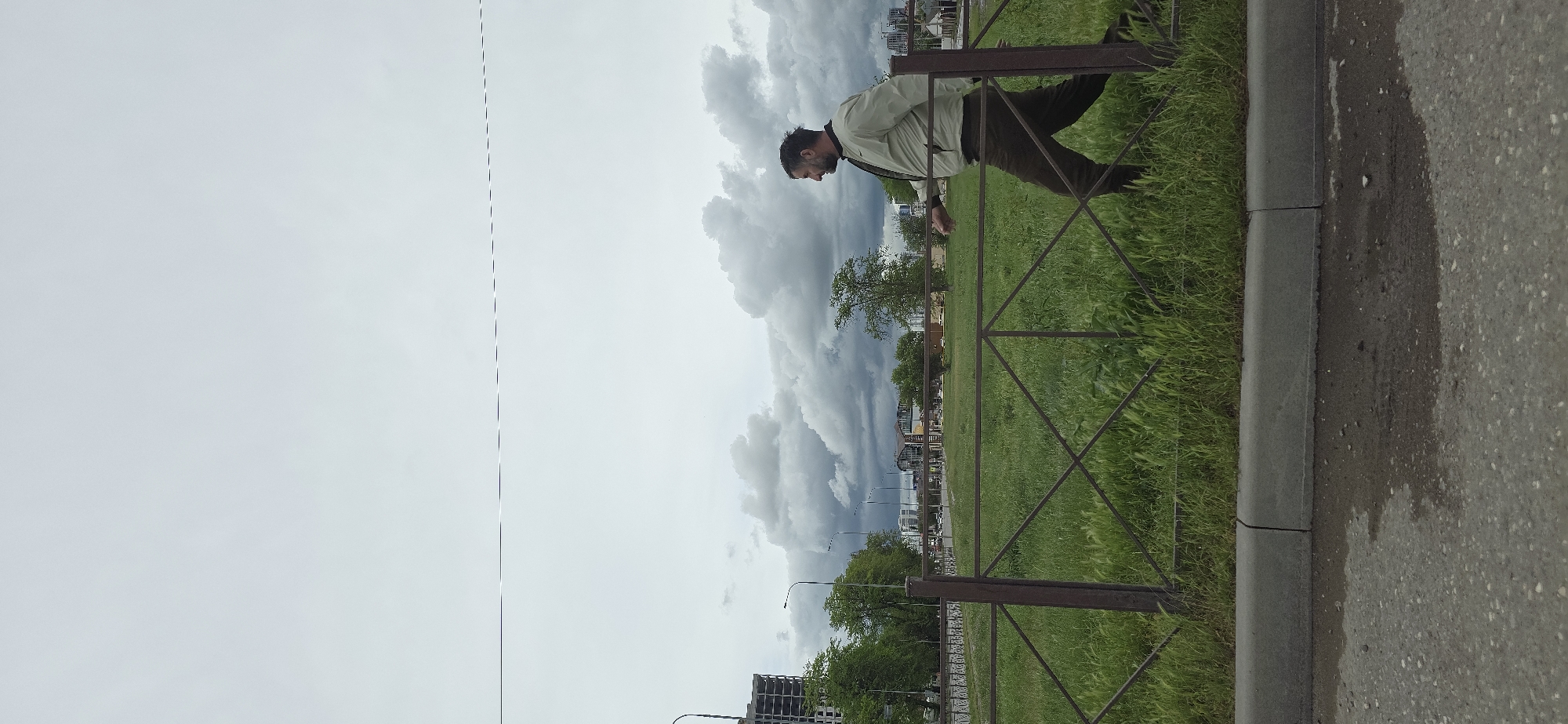 A man is standing by a metal fence in a park-like area. He is wearing a light-colored jacket and dark trousers. Behind him is a cloudy sky, trees, and buildings in the distance. The fence separates the grassy area from a sidewalk and street. A puddle of water is visible on the edge of the sidewalk. The location is in the city of Unknown, Unknown.