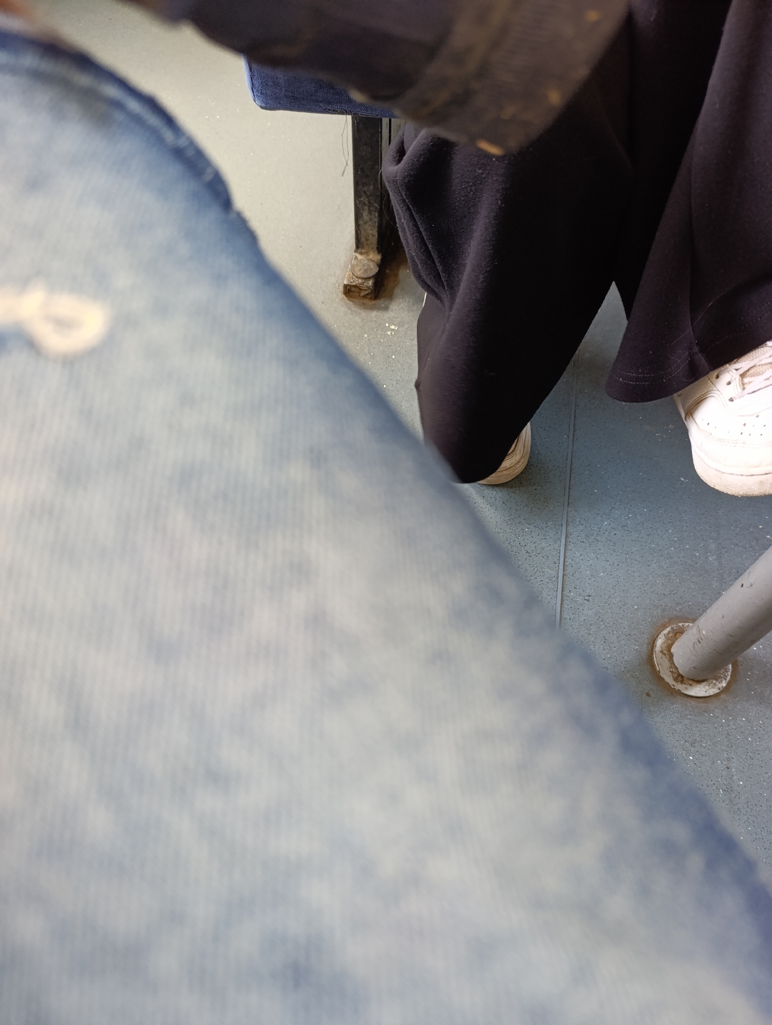 The image presents a close-up, first-person perspective of an indoor setting, likely a classroom, office, or cafeteria, in Stambolovo, Bulgaria. In the blurred foreground, a person's light blue jeans are visible, angled across the bottom left of the frame. In the middle ground, another individual is partially seen, wearing black trousers or sweatpants and white sneakers. Their legs and feet are positioned on a light blue or gray floor, which appears to be a linoleum or tiled surface with a visible seam running through it. The floor is speckled with small white particles, possibly dust or debris.
Two metal furniture legs are present: a dark, possibly black, square-profile leg with rust or dirt at its base is visible on the left, and a lighter gray, round-profile pole with a circular base plate, also showing signs of wear and rust, is on the right. The white sneakers appear worn, contributing to a sense of everyday use. No specific activity is discernible beyond the presence of individuals, presumably seated, and the general appearance of the floor and furniture suggests a functional, possibly institutional, environment. There is no visible text in the image.
