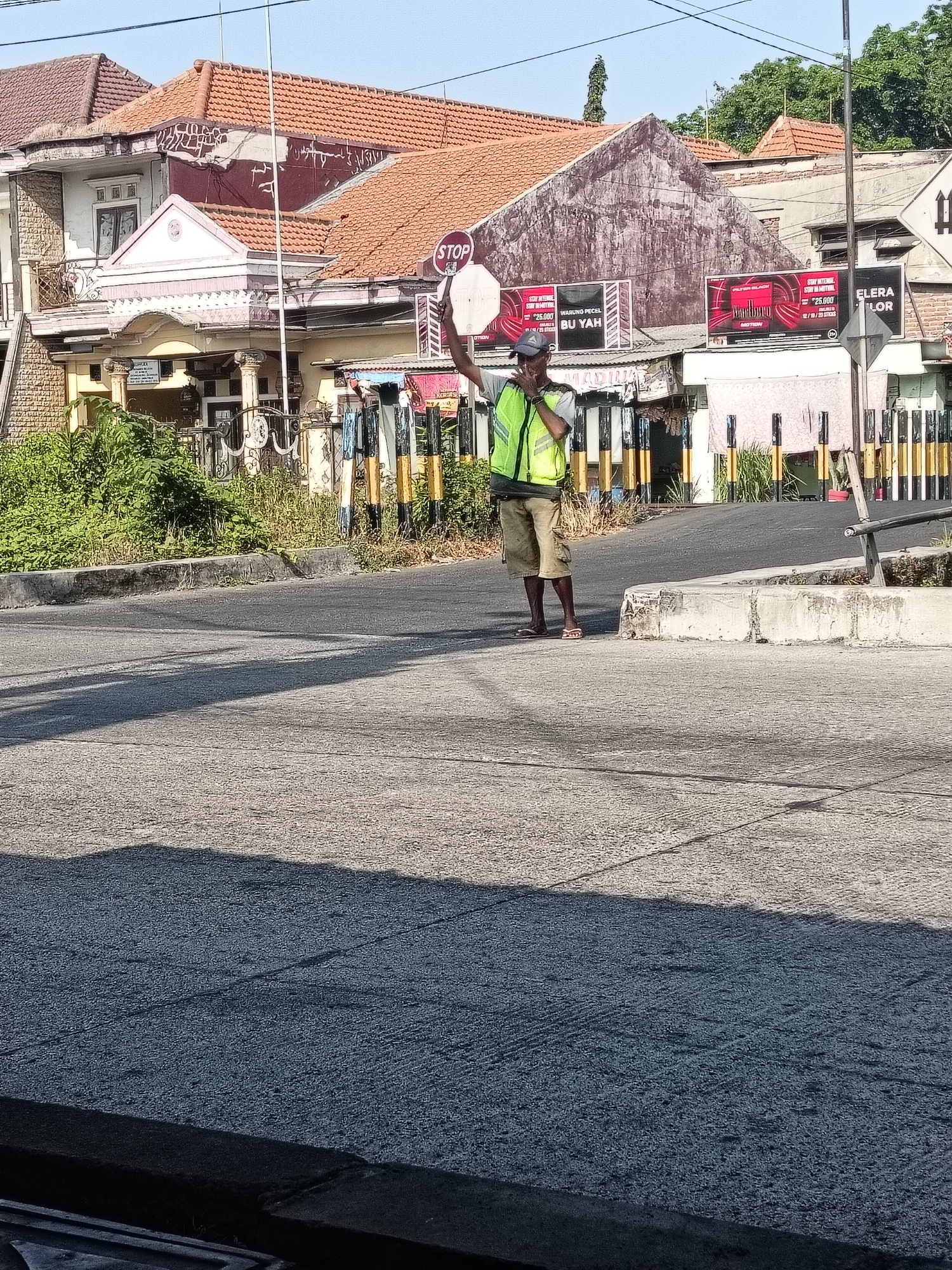 The image depicts a traffic controller in Gedangan, Indonesia, holding up a round "STOP" sign. He is wearing a bright green reflective vest over a light-colored t-shirt and khaki shorts, with flip-flops on his feet. The man is standing on an asphalt road, with houses and some billboards in the background. The houses have red tile roofs, typical of the region. The billboards appear to advertise local businesses. A concrete curb is visible in the foreground, casting a shadow on the road. The overall lighting suggests it is a sunny day. The scene appears to be a street intersection or a point where traffic control is necessary.
