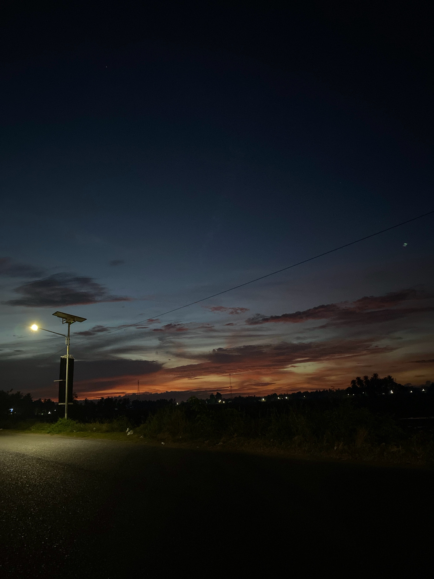 The image depicts a twilight scene on a rural road in Cotgapu, Indonesia. The sky is a deep blue transitioning to fiery oranges and pinks near the horizon, with scattered clouds. A solar-powered street lamp with its light on is positioned to the left of the frame. A power line stretches diagonally across the sky. The road in the foreground is asphalt and appears wet, reflecting the faint light. A dark silhouette of trees and vegetation lines the horizon, with some distant lights suggesting a village or settlement. There are no people visible in the image. The overall atmosphere is serene and quiet, characteristic of a rural evening.