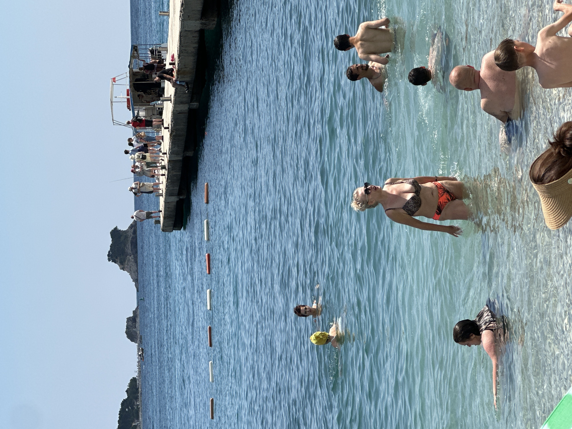 The image shows a sunny day at a beach in Budva, Montenegro. In the foreground, several people are in the shallow water. A woman in a bikini is standing, looking to the left. Behind her, a man and a boy are partially submerged. Further back, a man is swimming, and two people in yellow and black caps are also in the water. In the mid-ground, a pier juts out into the sea with several people standing on it, some of whom are near a small boat. The water is clear and blue, with a few buoys floating on the surface. In the background, there are rocky islands covered in green foliage and the coastline with buildings. The sky is clear and blue.