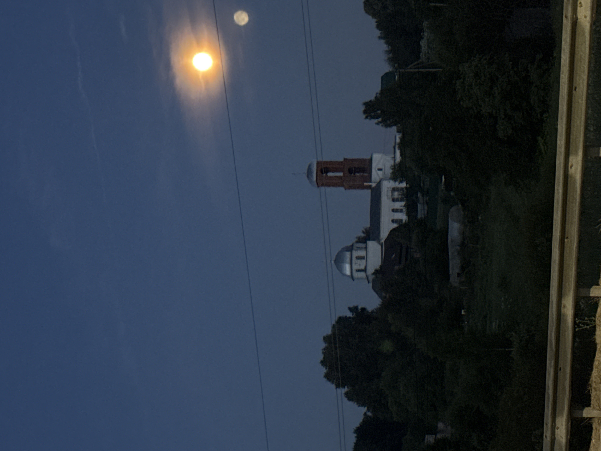 The image depicts a twilight scene in Svatkovo, Russia, featuring a church complex surrounded by trees. Two celestial bodies, likely the moon and a smaller satellite or planet, are visible in the deep blue sky, with one illuminated by the setting sun and the other faintly visible. A cluster of buildings, including a church with a silver dome and a brick bell tower, is nestled among the trees in the mid-ground. The foreground is dominated by a wooden barrier, suggesting the vantage point is from a road or elevated area. The overall atmosphere is calm and serene, with the fading light casting long shadows and creating a sense of depth.