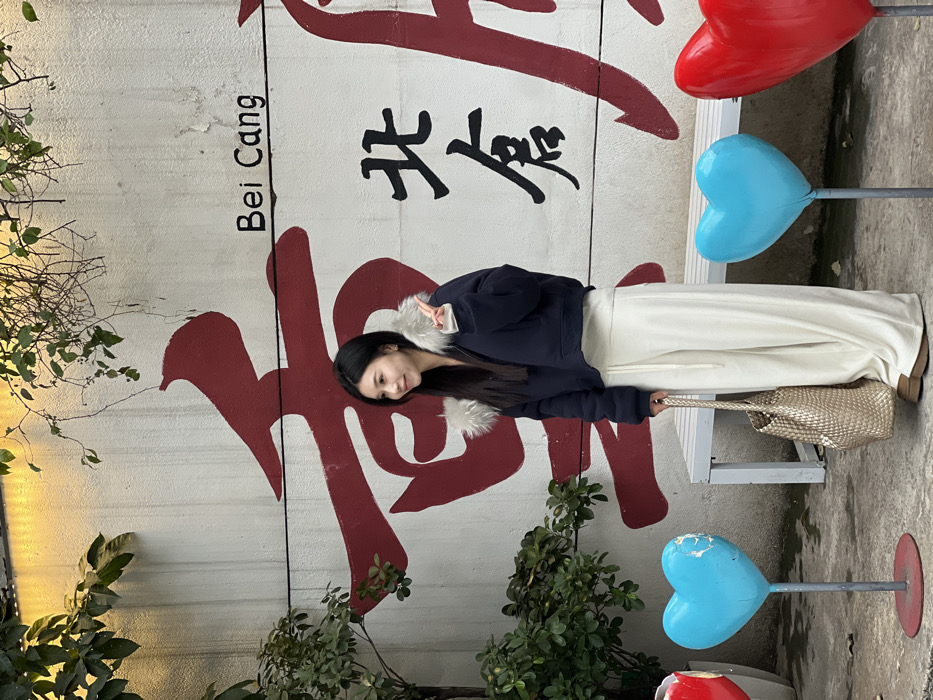An East Asian female subject is depicted standing outdoors against a white wall in Bei Cang, Liangjiang New Area, Chongqing, China. She has long, dark hair and is smiling, looking towards the viewer's left. Her left hand is raised, forming a peace sign. She is dressed in a dark navy cropped puffer jacket with light-colored fur trim on the hood and cuffs, cream-colored wide-leg pants with a drawstring waist, and light brown platform shoes. She holds a woven gold/beige handbag in her right hand.
The white wall behind her features large red Chinese characters, with "Bei Cang" written in black English text above them, and "北倉" (Bei Cang) in black Chinese characters further to the right. Green foliage is visible along the top and left edges of the wall. A white slatted bench is positioned against the wall behind the subject. On the ground in the foreground, three large heart-shaped sculptures on poles are visible: a partial red heart on the far left, a light blue heart with some wear to the left of the subject, and a red heart to her right. The scene is illuminated by natural daylight.