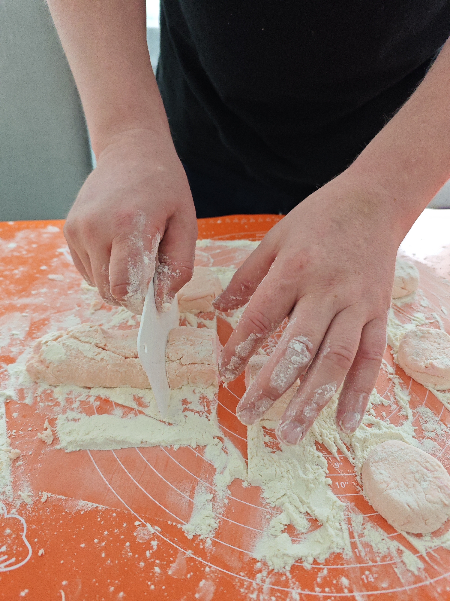 A person wearing a black shirt is cutting dough with a white plastic scraper on an orange silicone baking mat covered in white flour. The person's hands are also covered in flour. The mat has white measurement markings. The location is assumed to be Markovskii, Russia.
