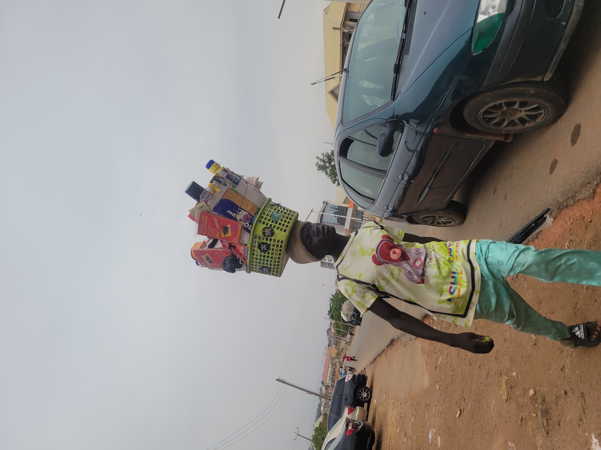 The image displays a street scene in what appears to be a town or city, likely in Jos, Nigeria. A young Black male, positioned centrally, is depicted standing or walking along the side of a road. He is engaged in street vending, carrying a large, green plastic basket laden with various packaged goods balanced on his head, which is cushioned by a straw hat.
The contents of the basket include numerous items such as bottles (one with a blue cap), cartons, and other packaged products. Visible text on some of these items includes "BEST," "SUPER," "THIS IS YOUR BADGE," "P_PER," and "S_AVOURY JU_CE."
The man wears a light-colored, possibly cream or white, short-sleeved shirt adorned with a graphic of a teddy bear wearing headphones, light blue or green pants, and black sandals. The "adidas" logo is clearly visible on one of his sandals. His right arm is slightly bent, and he holds a small green object in his hand. His expression is neutral, reflecting focus.
To the right of the man, a dark green car, possibly a sedan, is parked. Its front section, including the windshield, headlights, and front tires, is visible. Further down the road, another dark-colored car is also parked. The road consists of a dirt-colored shoulder on the left transitioning to a paved surface with some visible stains or marks.
The scene is set outdoors under an overcast sky, indicating daytime. In the background, low-rise buildings line the street, accompanied by utility poles and overhead wires, contributing to the urban or semi-urban setting.