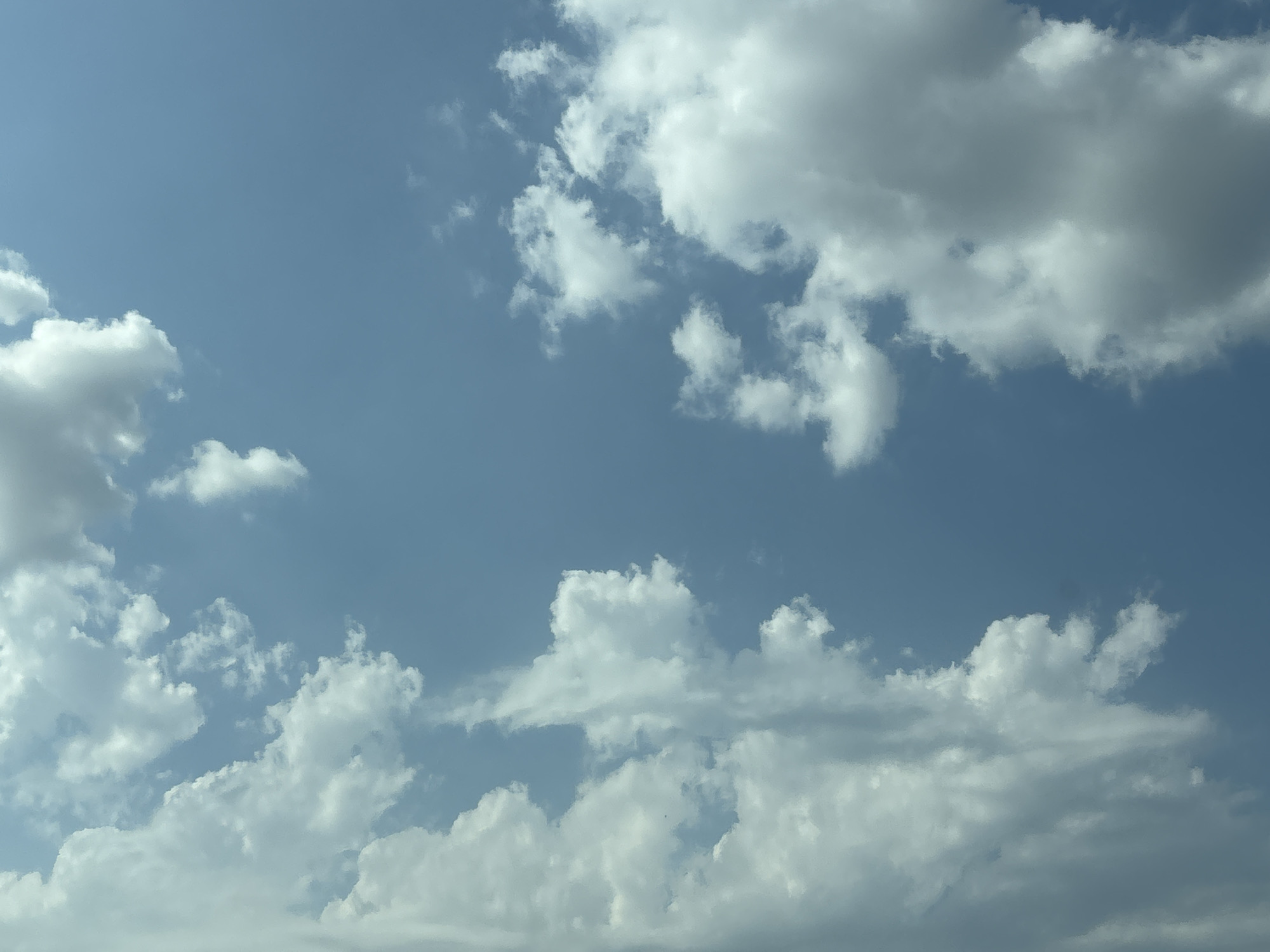 The image is a photograph of a cloudy sky over a city. The sky is a light blue, with white and gray clouds scattered across it. Some of the clouds are large and puffy, while others are smaller and wispy. The image appears to have been taken during the daytime, as the sun is not visible but the sky is bright. The clouds are casting shadows on each other, indicating the presence of sunlight. The bottom part of the image shows a hint of city buildings, though they are out of focus and not the primary subject. The overall impression is one of a typical cloudy day, with the sky being the dominant feature. The image does not contain any people or identifiable landmarks, making it difficult to pinpoint an exact location beyond the general description of a city.